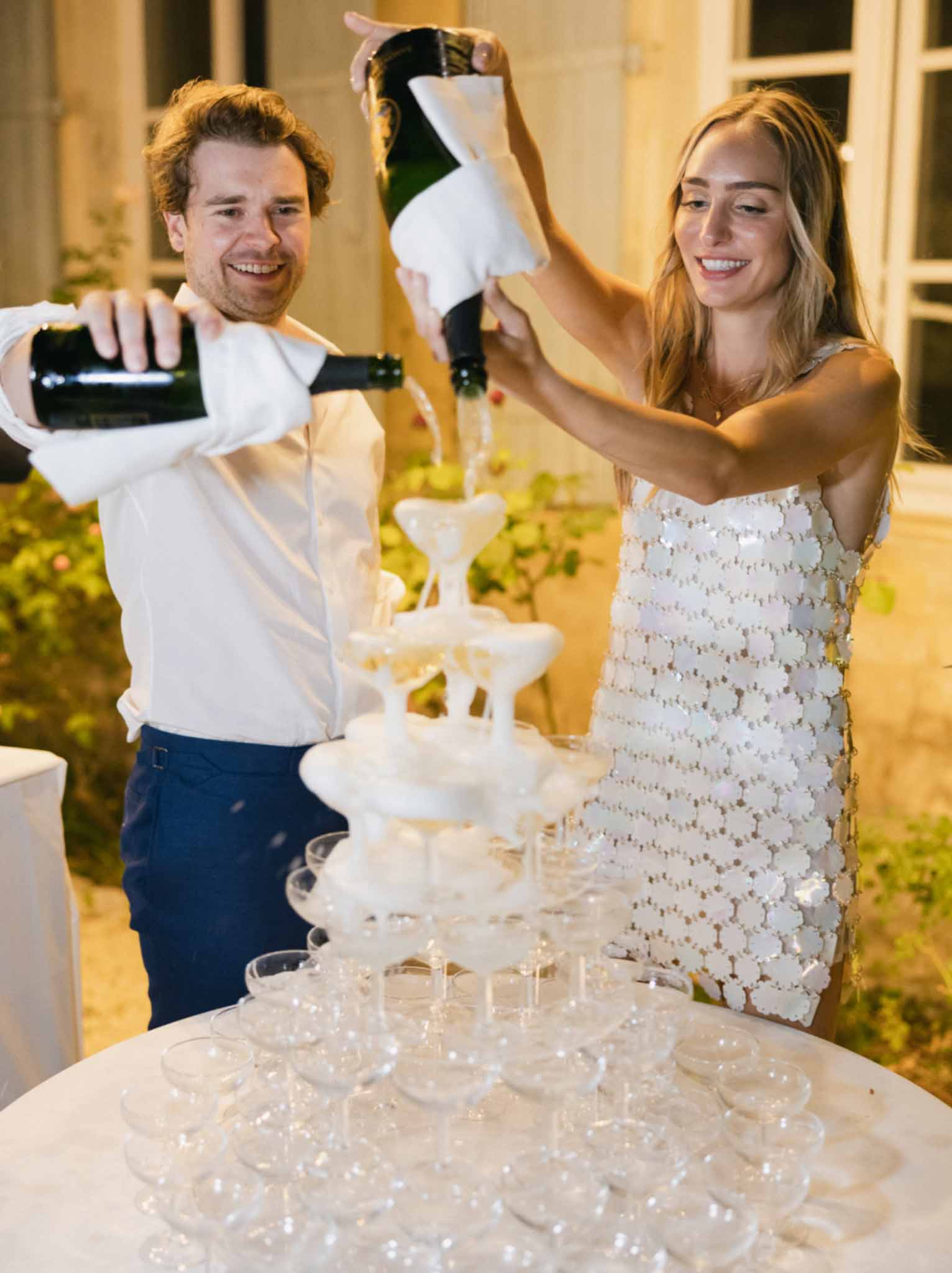 Bride and groom pouring champagne into a tiered coupe tower inside an ornate ballroom