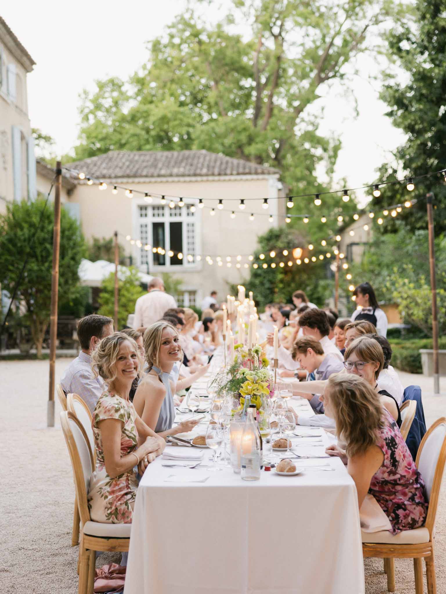 Guests seated at long candlelit dinner table with yellow and green floral centerpieces under string lights in courtyard