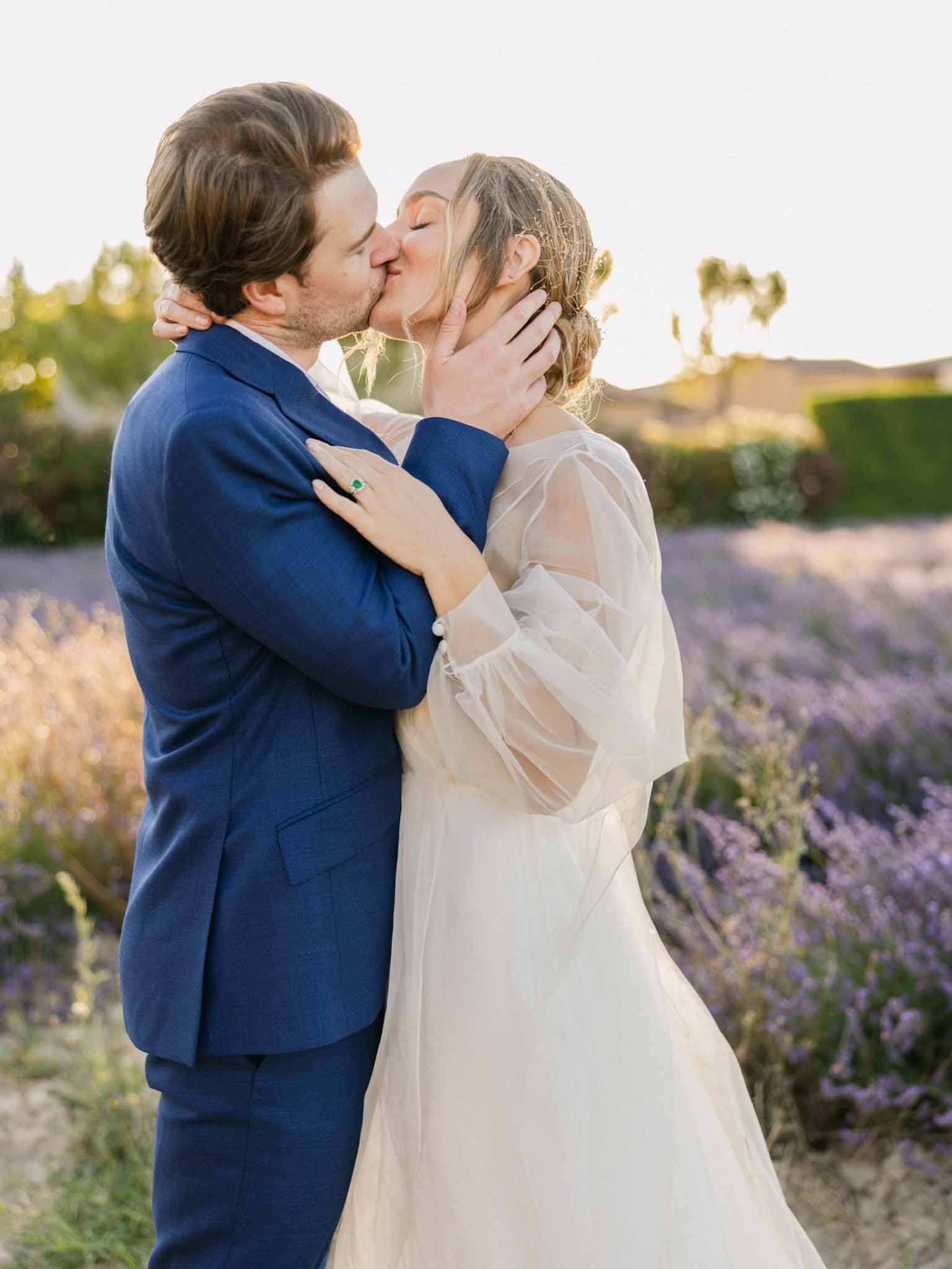 Bride and groom kissing in a lavender field at golden hour; bride in ivory tulle gown, groom in navy suit.