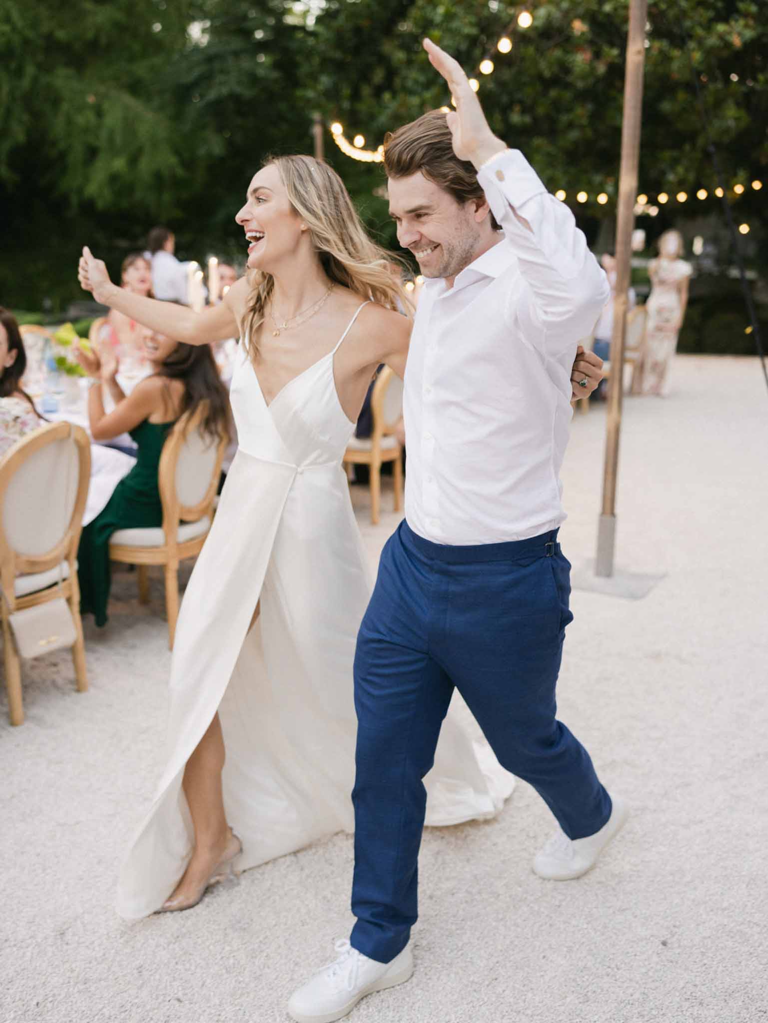 Bride and groom dancing joyfully at evening reception with string lights and wooden chairs