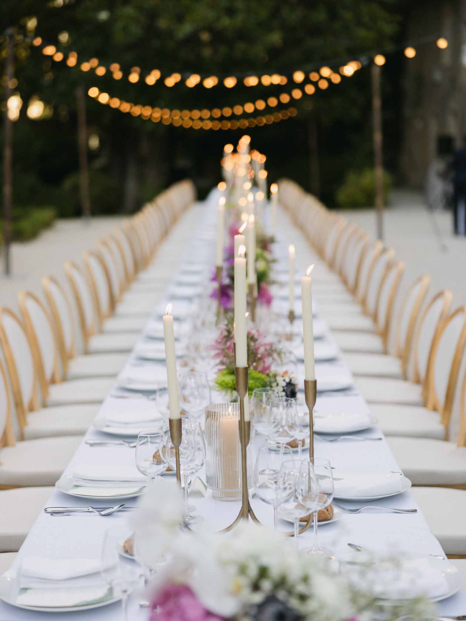 Long banquet table set for outdoor reception with brass candlesticks, white and purple florals, and string lights overhead