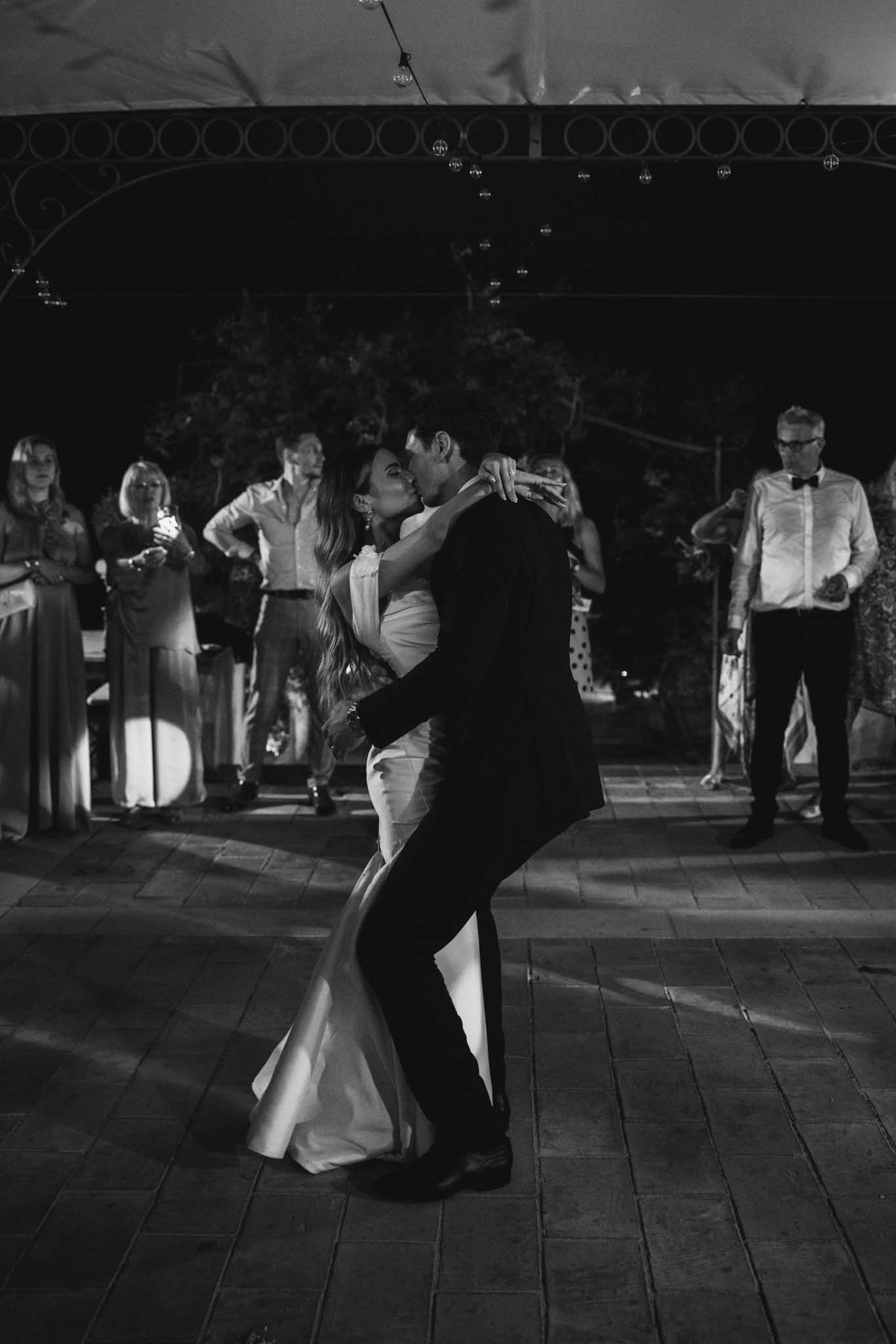 First dance under covered venue, black and white, guests watching from perimeter, pendant lights above tiled floor