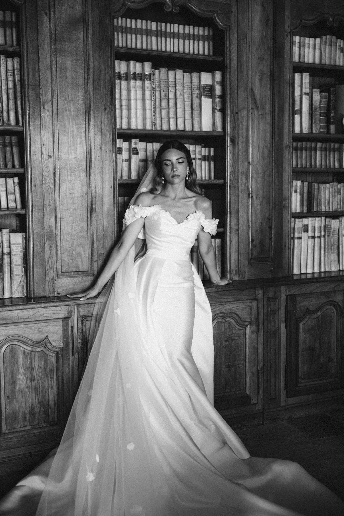 Black and white bridal portrait of bride in off-shoulder gown standing in chateau library