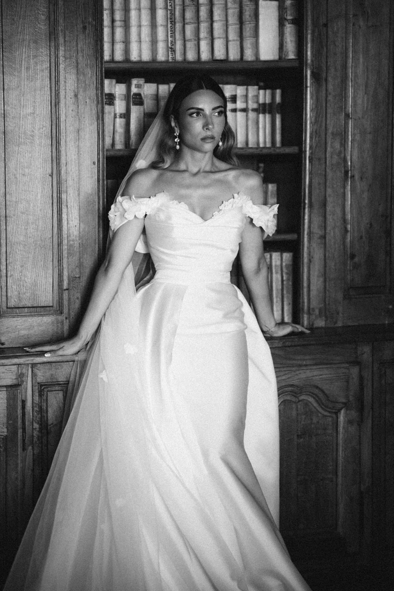 Black-and-white bridal portrait in off-shoulder ruched dress with long veil against dark wooden bookcase