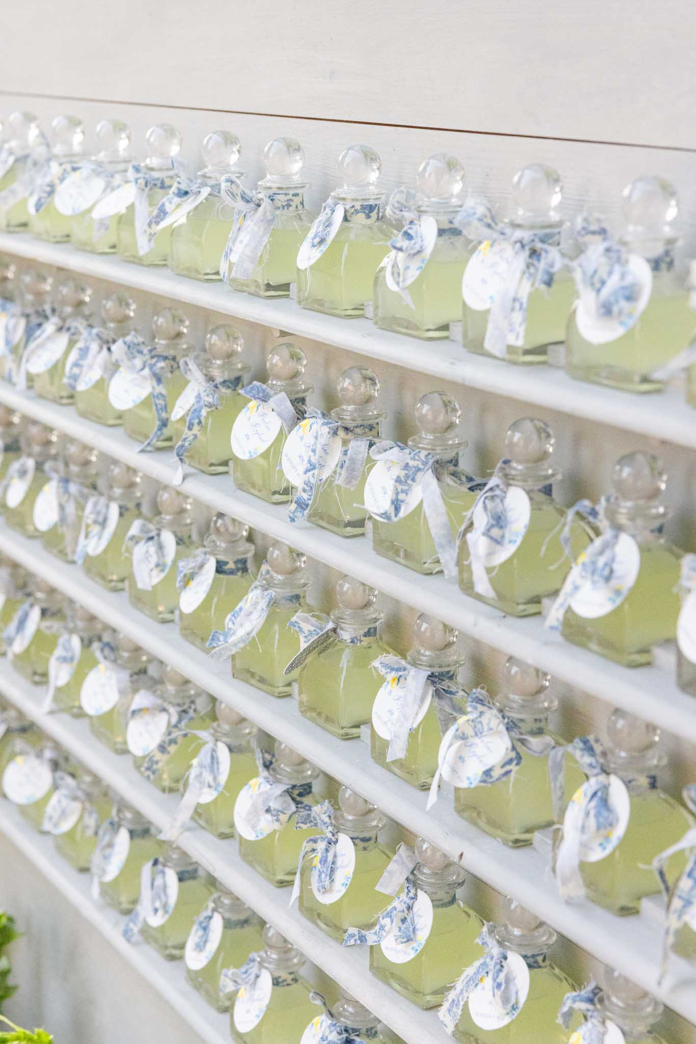Rows of small glass bottle wedding favors with spherical stoppers tied with blue and white floral ribbon on white shelving