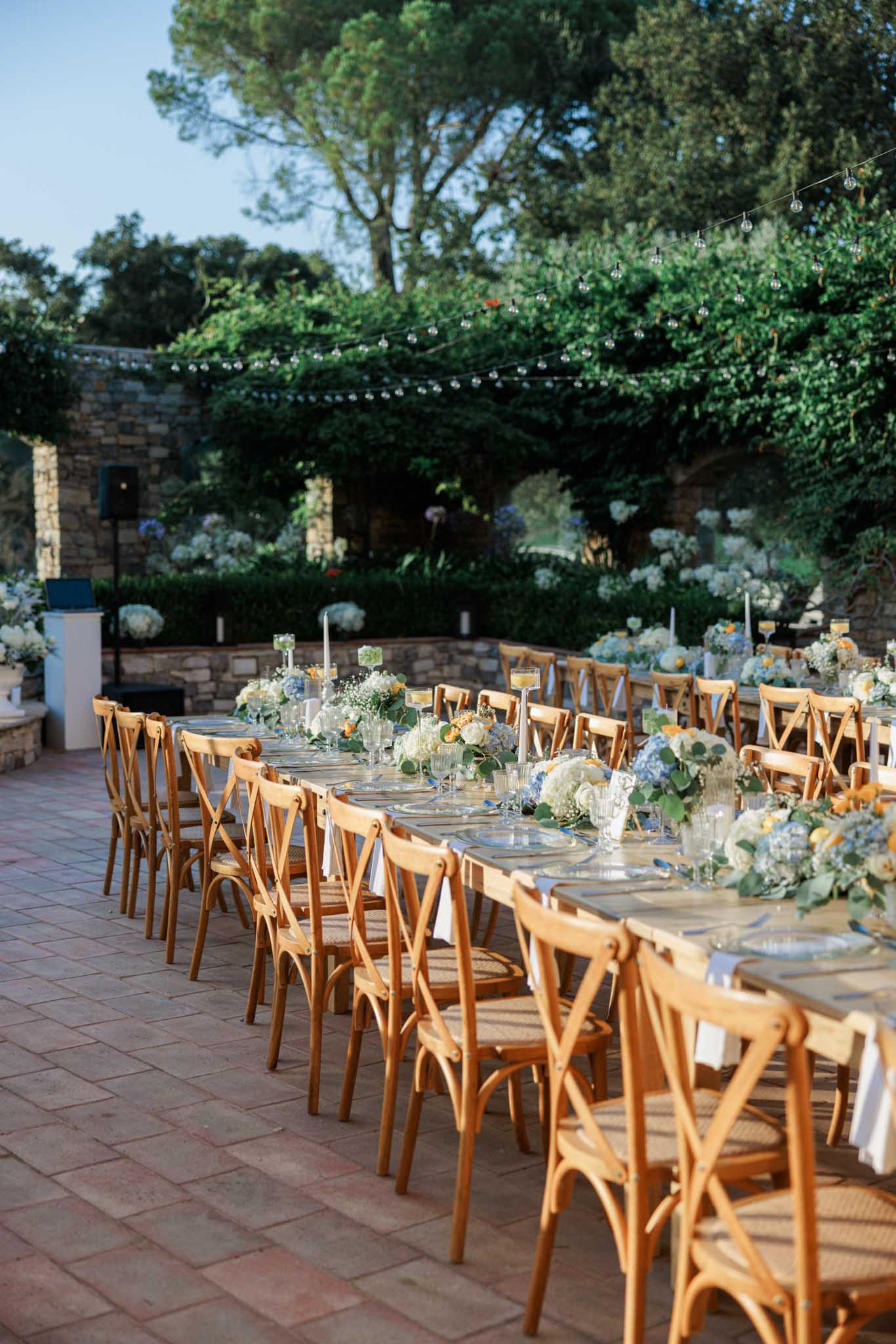 Outdoor reception tables on brick terrace with white hydrangeas, cream and yellow roses, and cross-back chairs