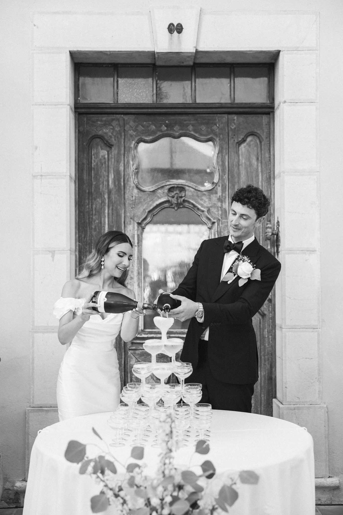 Bride and groom performing champagne tower ceremony in front of ornate wooden door, black and white photo