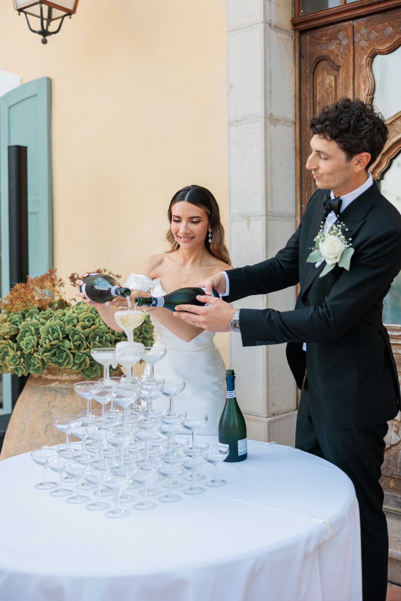 Groom pours champagne into tower of coupes while bride watches in Mediterranean courtyard with ochre walls