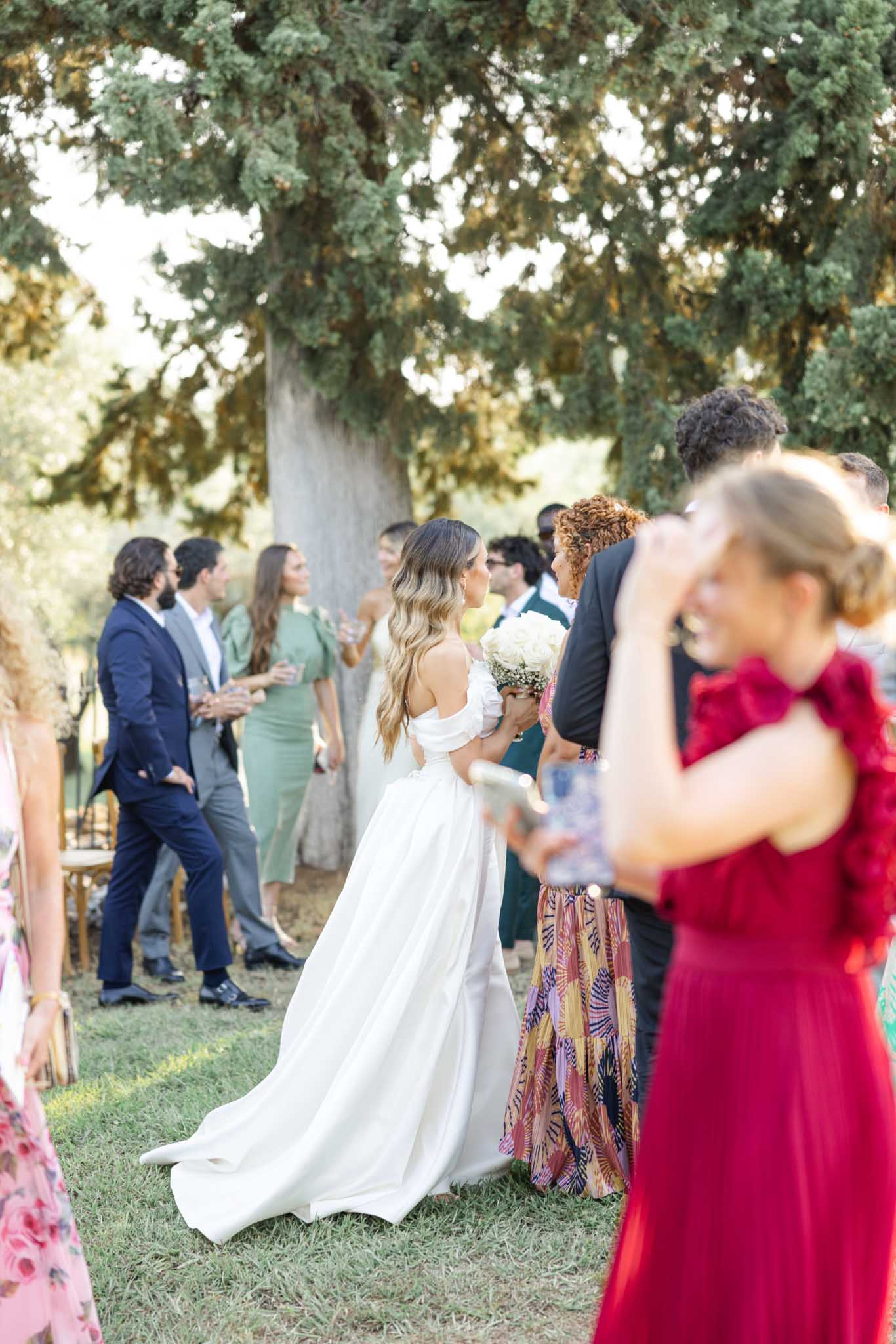 Bride mingling with guests during cocktail hour under dappled tree canopy