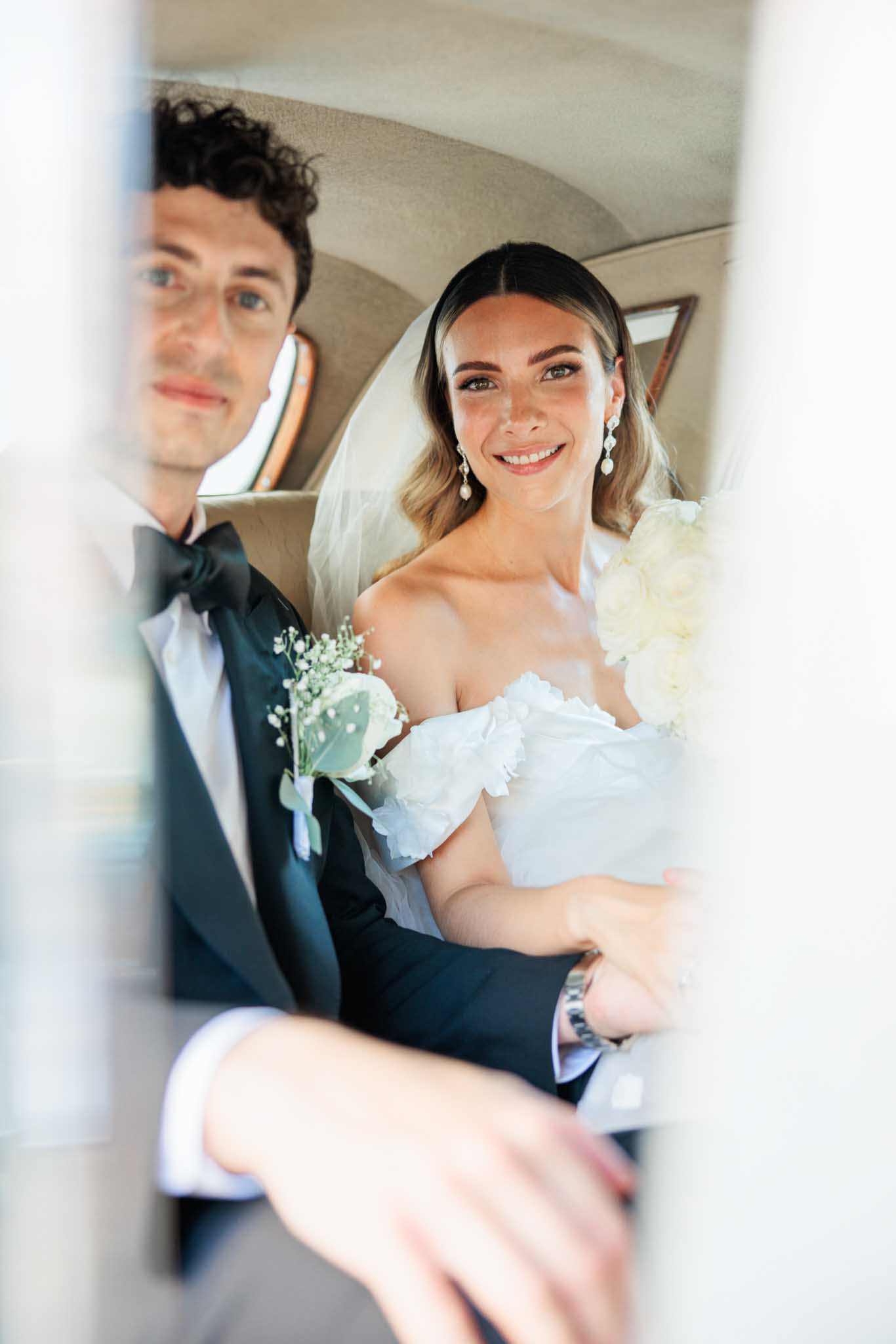 Bride and groom smiling through car window, bride in off-shoulder white dress, groom in black tuxedo