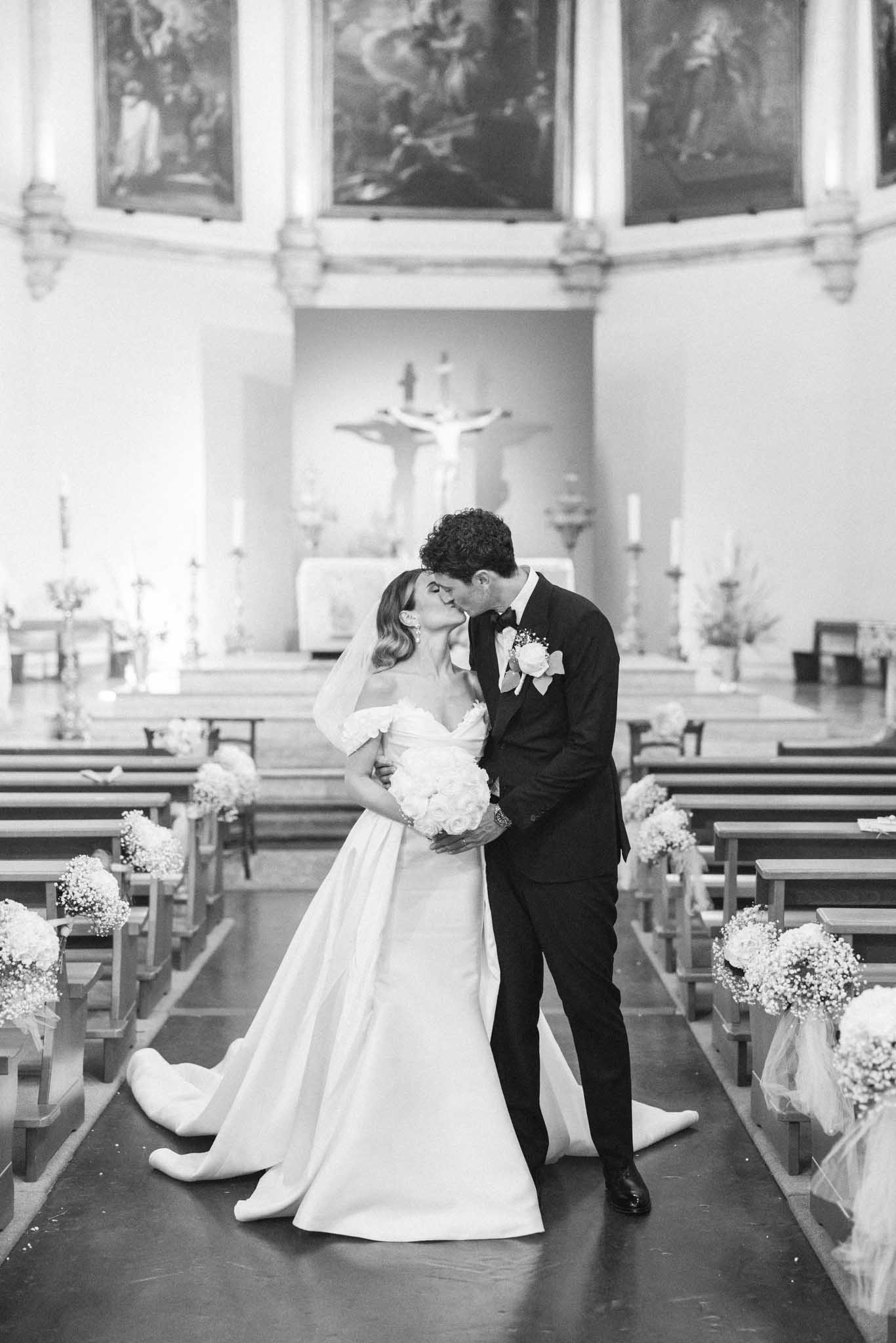 Bride and groom sharing first kiss at chapel ceremony with candles and pew florals, black and white photo