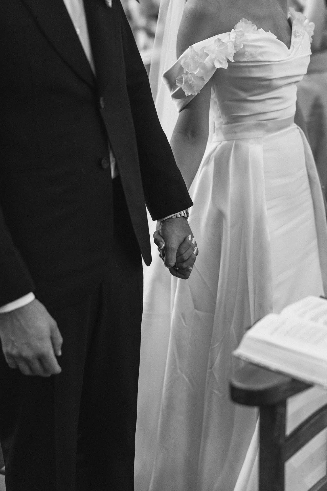 Black and white close-up of bride and groom holding hands during ceremony, groom's watch visible, officiant behind