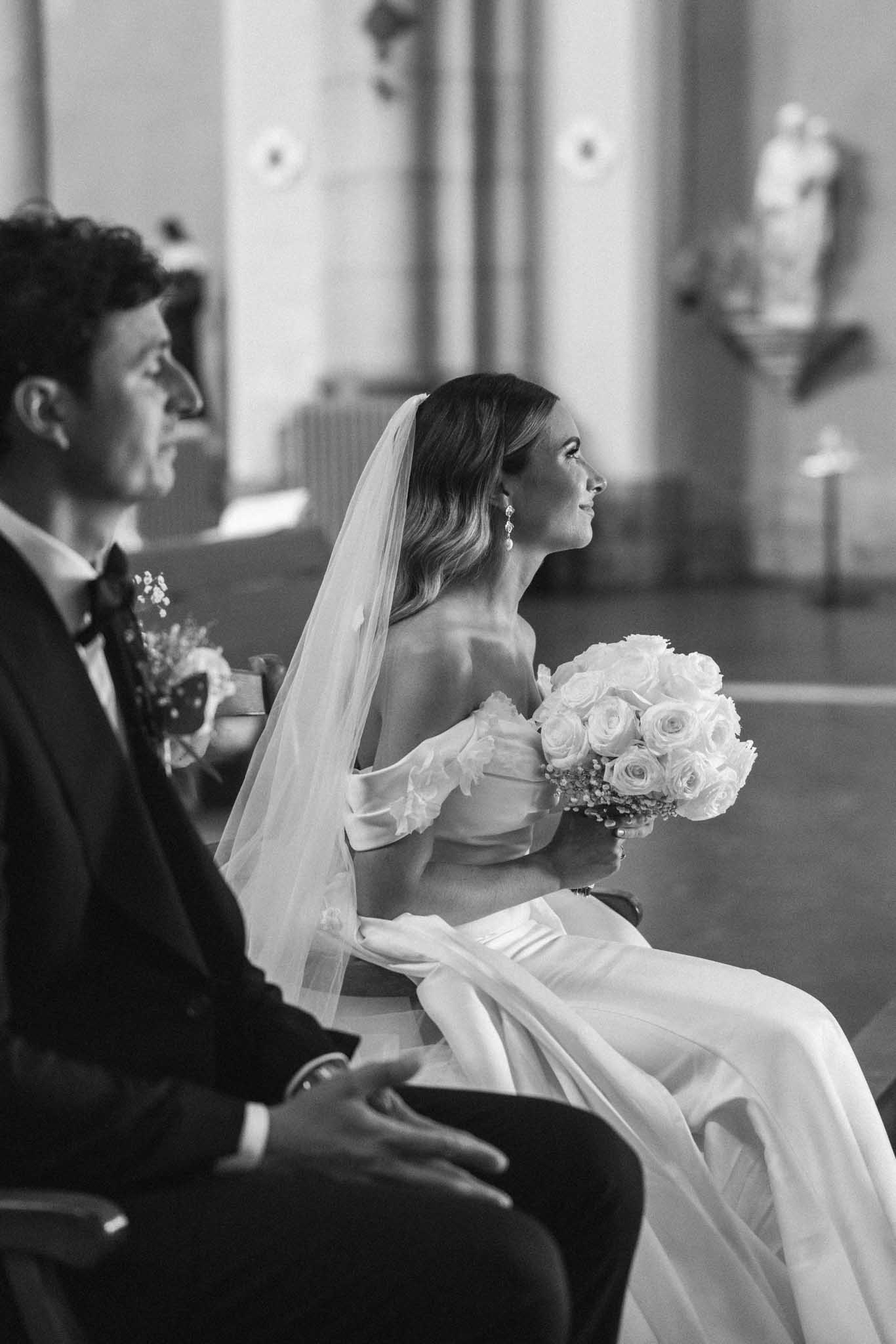 Black-and-white side profile of bride in strapless gown with veil and bouquet seated beside groom during indoor chapel ceremony