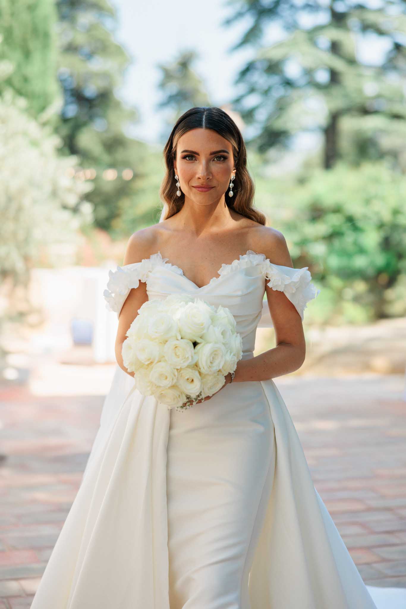 Bride in ivory off-shoulder ruffled gown holding round ivory garden rose bouquet on brick pathway