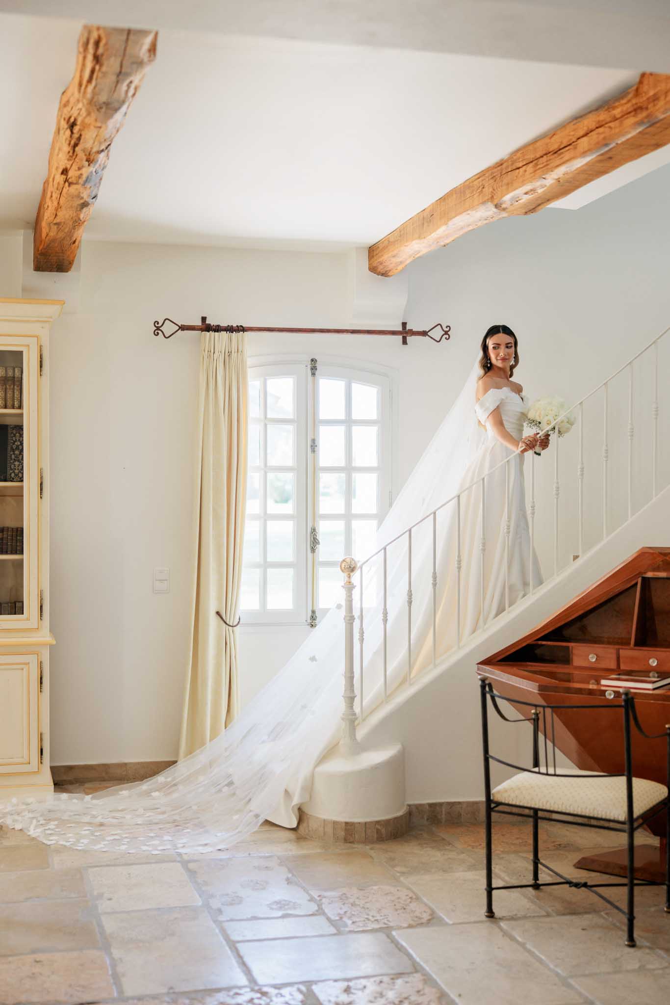 Bride in off-the-shoulder ivory gown poses on curved staircase with bouquet in rustic château interior