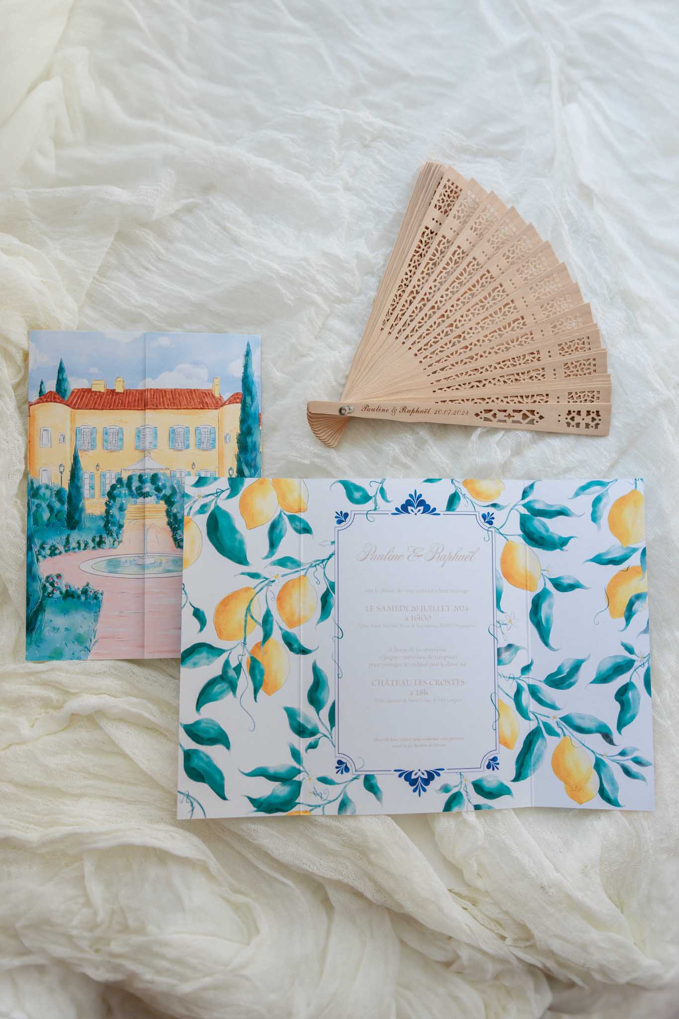 Flat lay on ivory tulle with Mediterranean villa invitation, lemon border design and laser-cut wooden fan