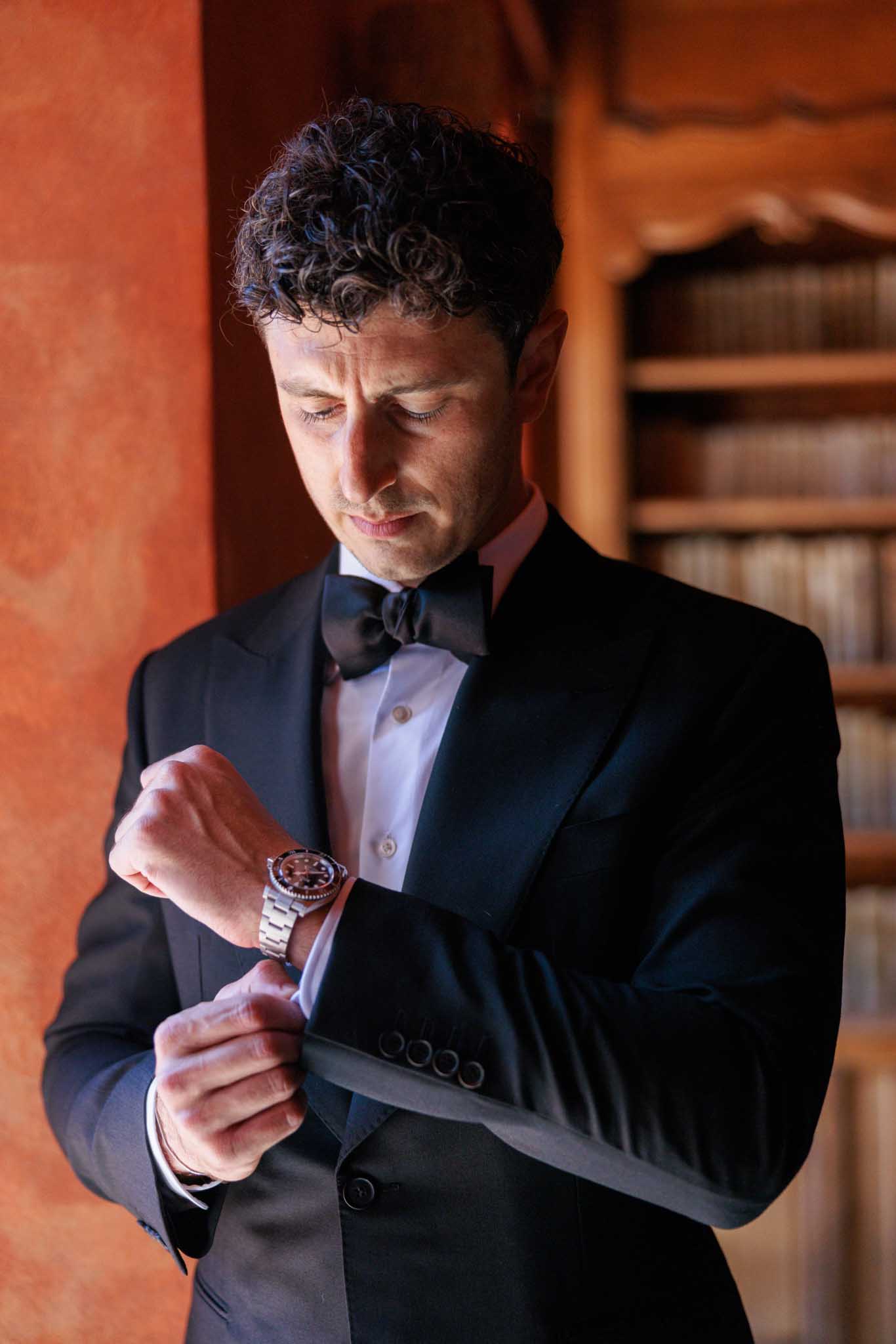 Groom in black tuxedo adjusting cufflink against terracotta-orange wall during wedding preparations