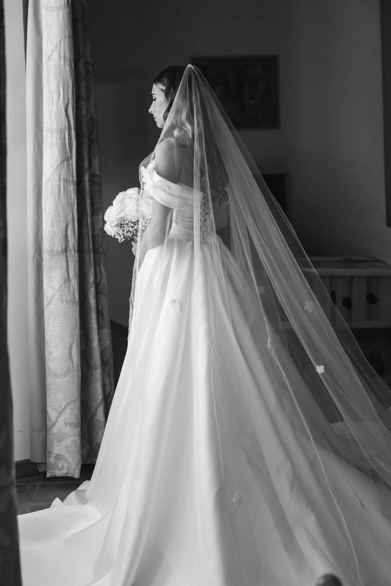 Black and white profile portrait of bride in strapless gown and cathedral veil holding bouquet near a window