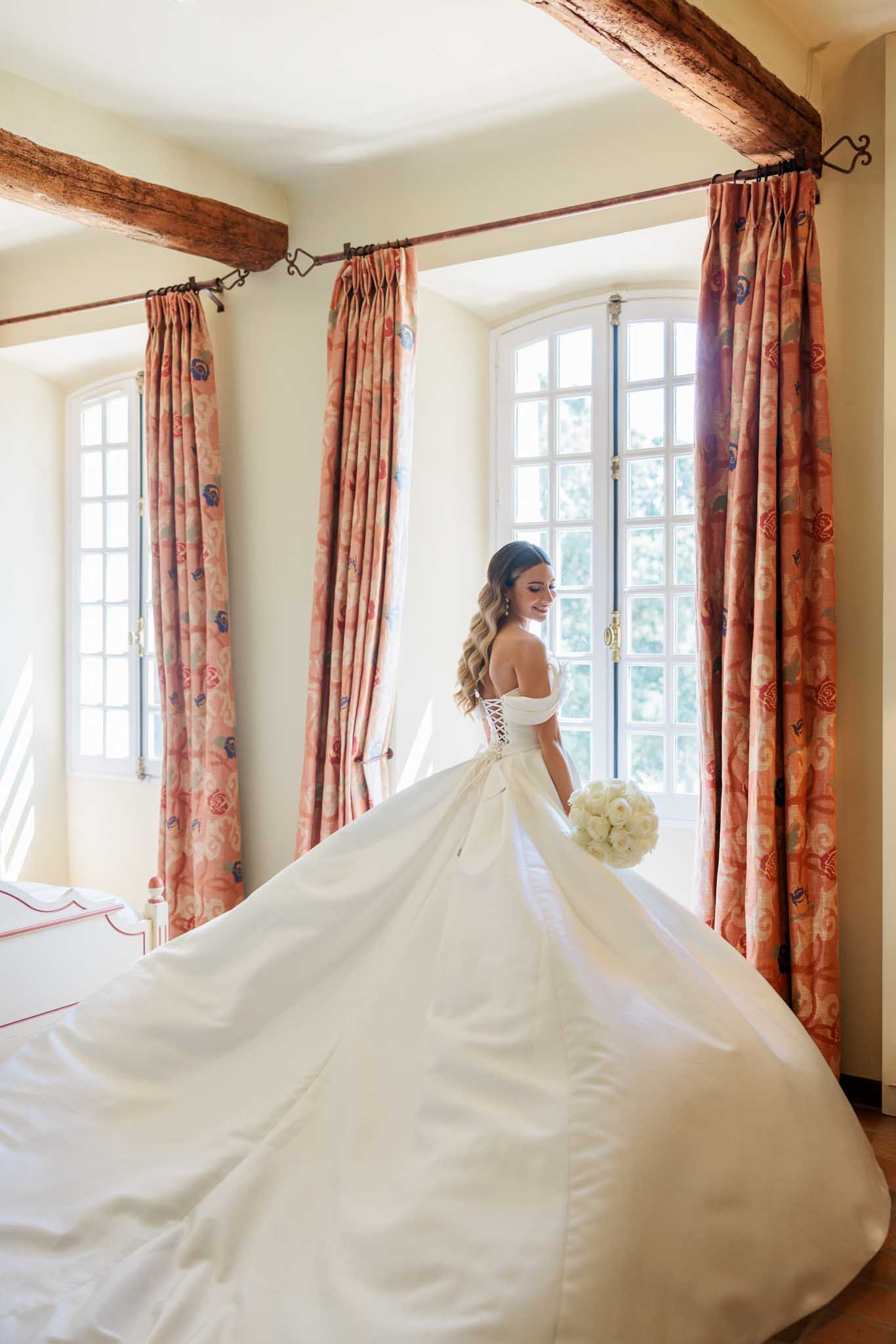 Bride in off-shoulder corset gown with flowing train holding white roses in classical room with French doors