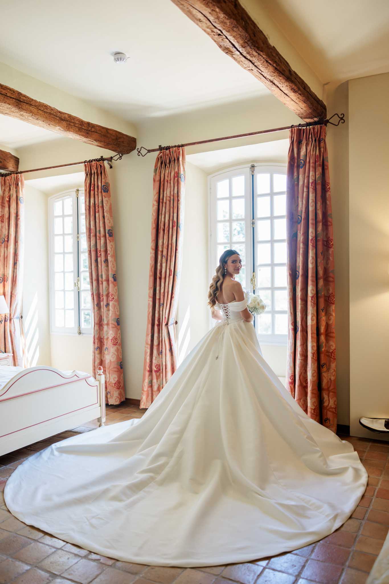 Bride in ivory off-shoulder ball gown holding bouquet in château bedroom with exposed wooden beams