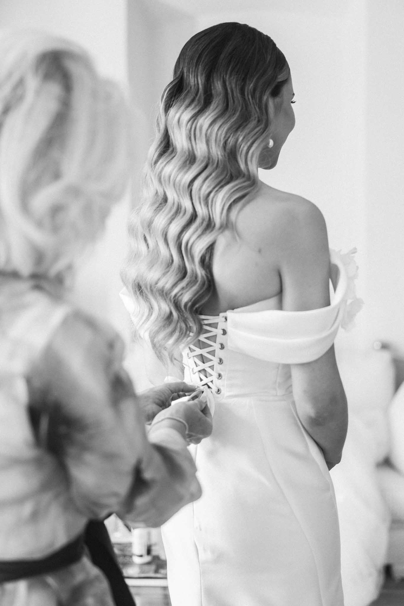 Black and white portrait of bride from behind in strapless corset-laced gown, waved hair, assistant partially visible