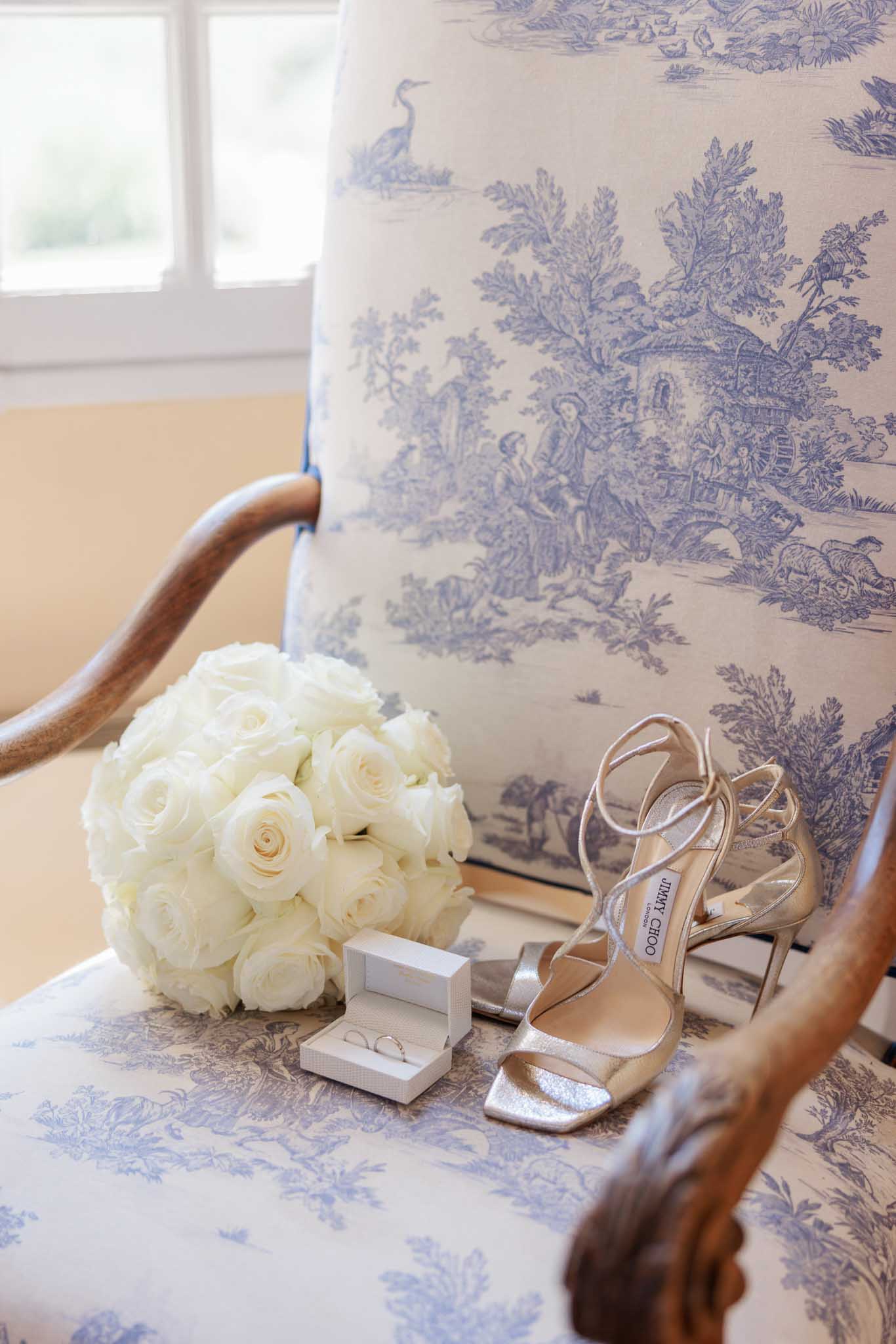 Bridal accessories on toile de Jouy chair including white rose bouquet, rose gold heeled sandals, and wedding rings