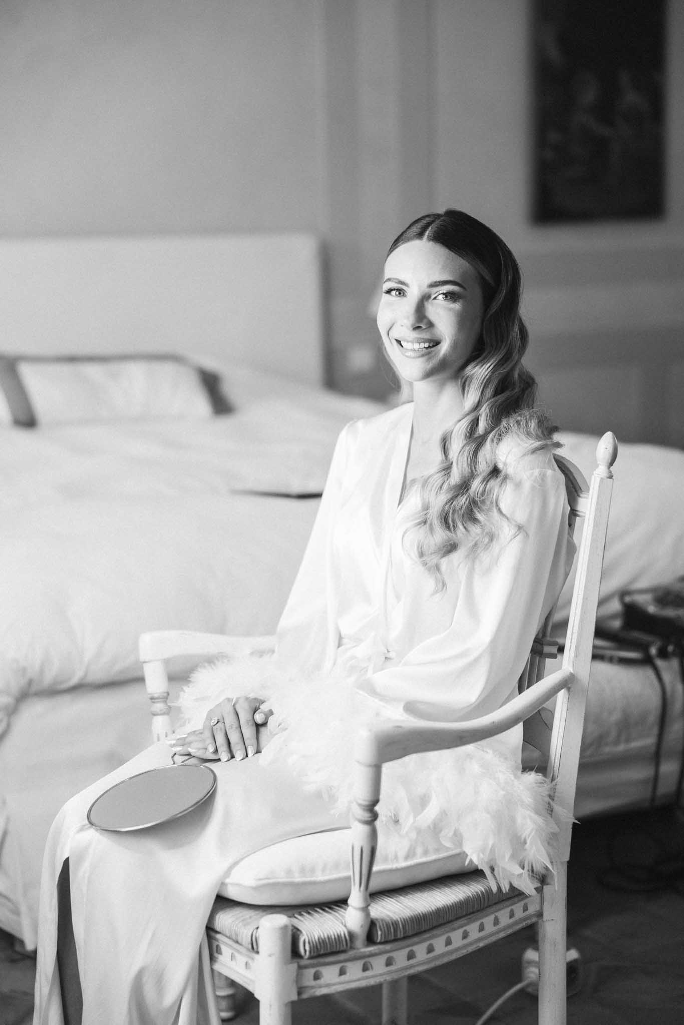 Black and white portrait of bride in white robe with wavy hair seated on a chair in a bridal suite during getting-ready preparations.