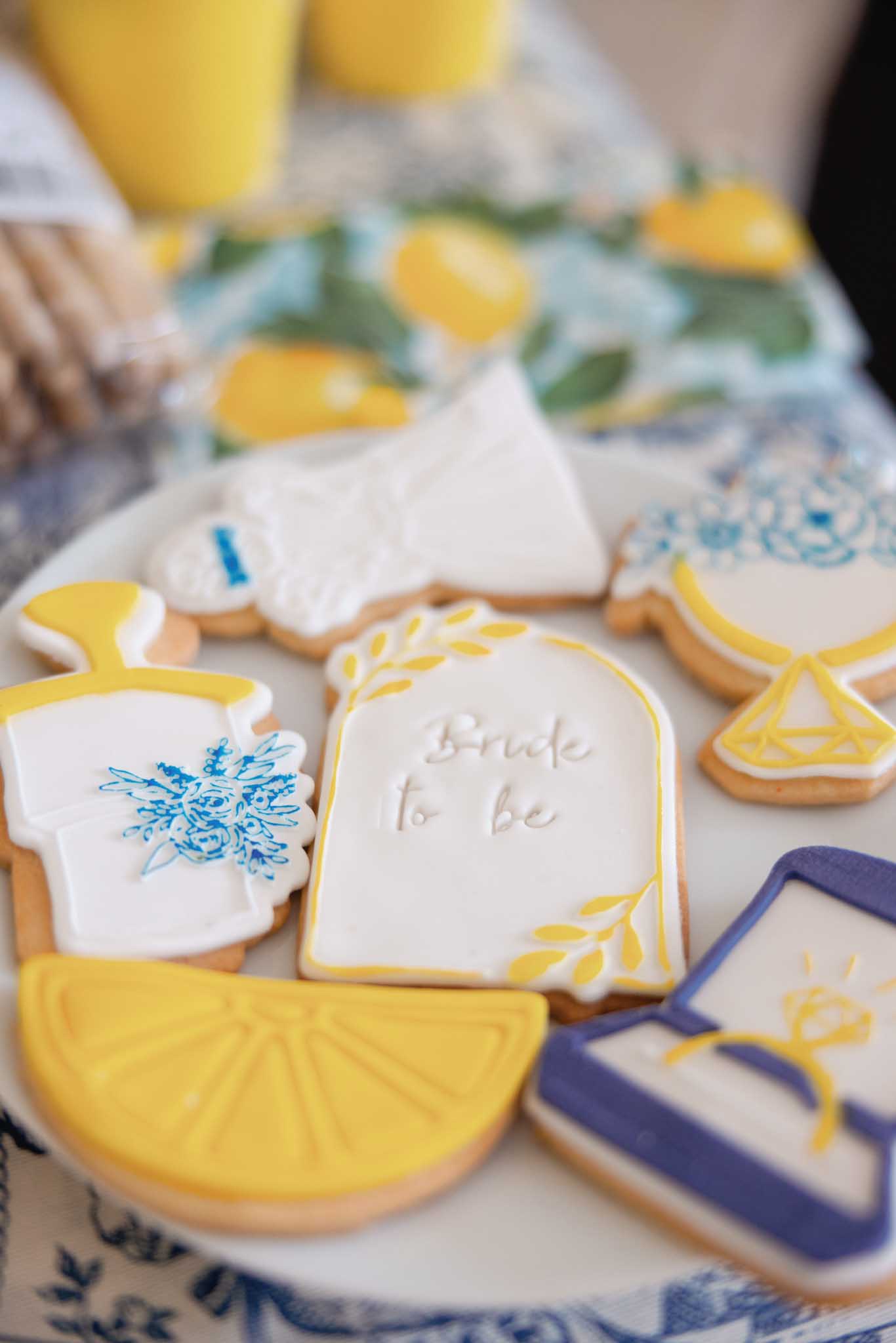 Decorated butter cookies in bride dress, lemon, and arch shapes with yellow and navy icing on patterned cloth
