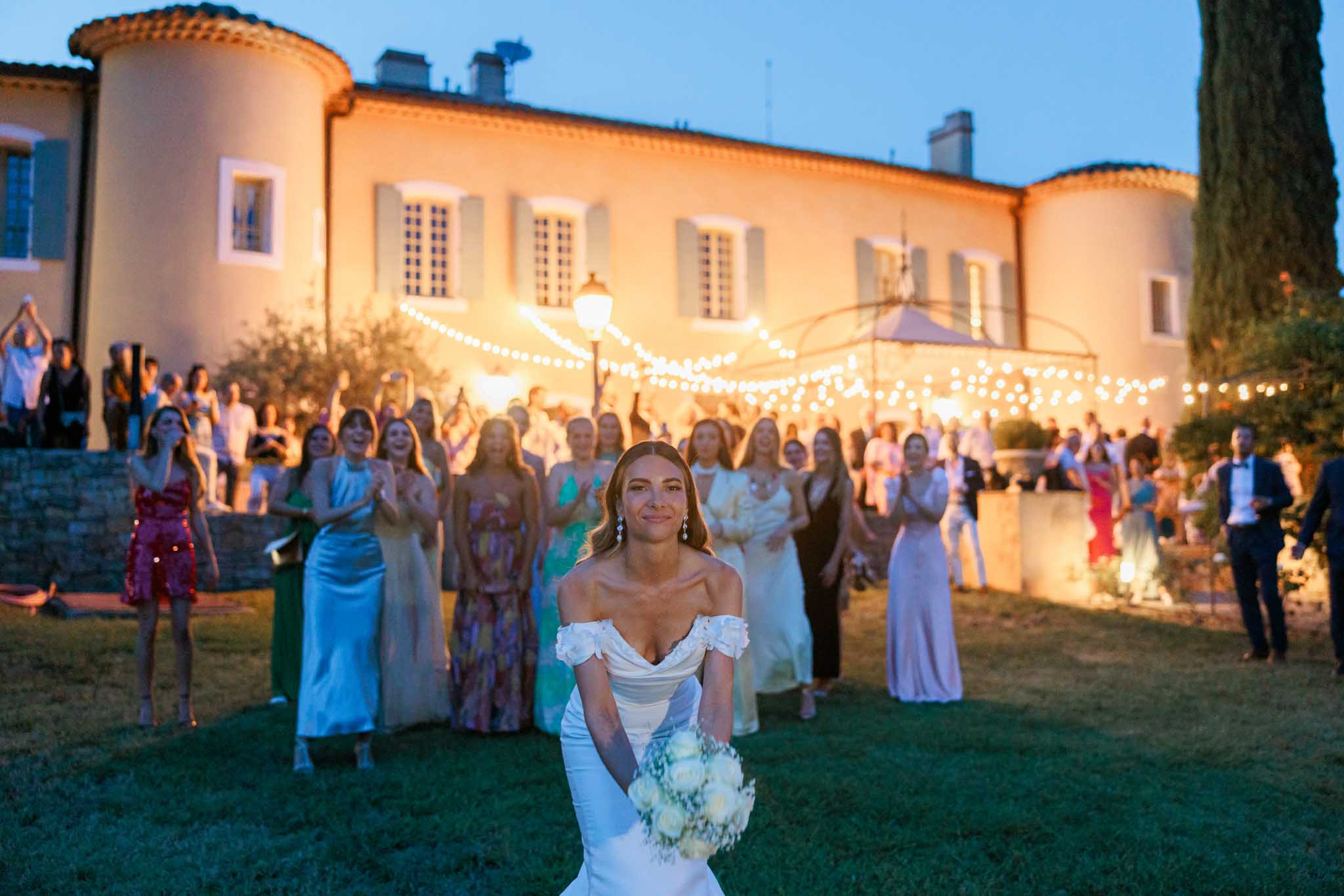 Bride in mermaid gown with guests and illuminated chateau facade with bistro lights at evening reception