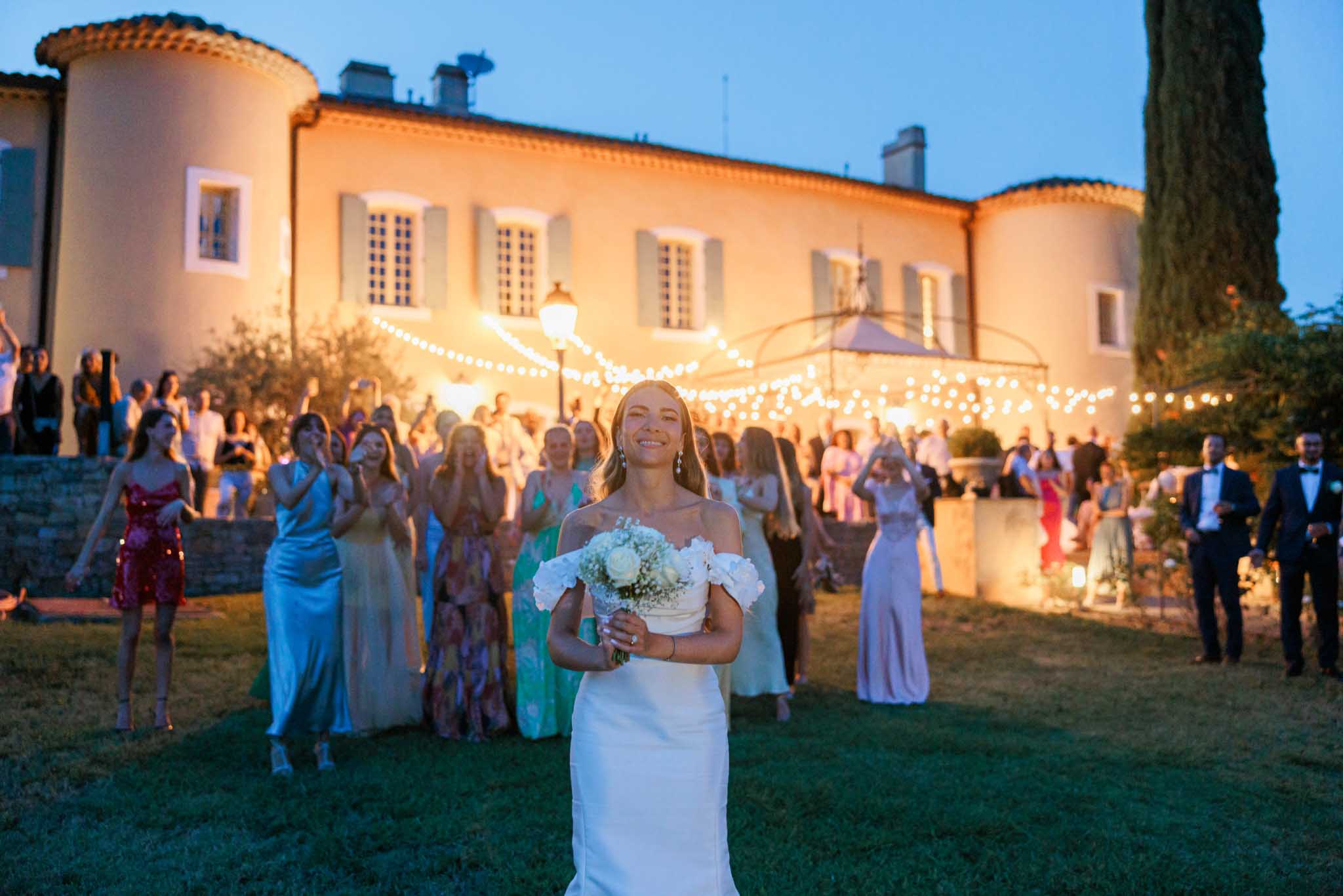 Bride holding bouquet in foreground with reception guests gathered on lawn before illuminated terracotta villa