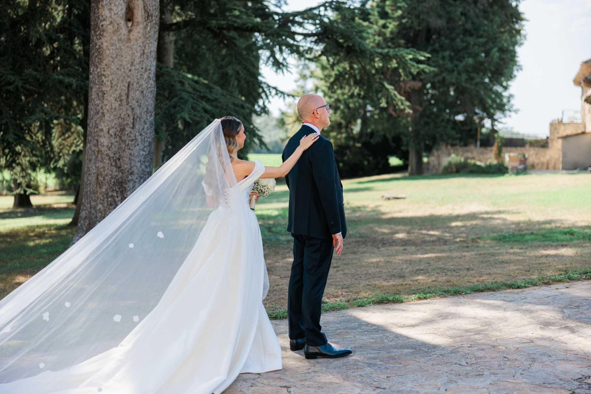 Bride in off-shoulder ivory dress with floral veil and groom in navy suit on stone path at chateau grounds