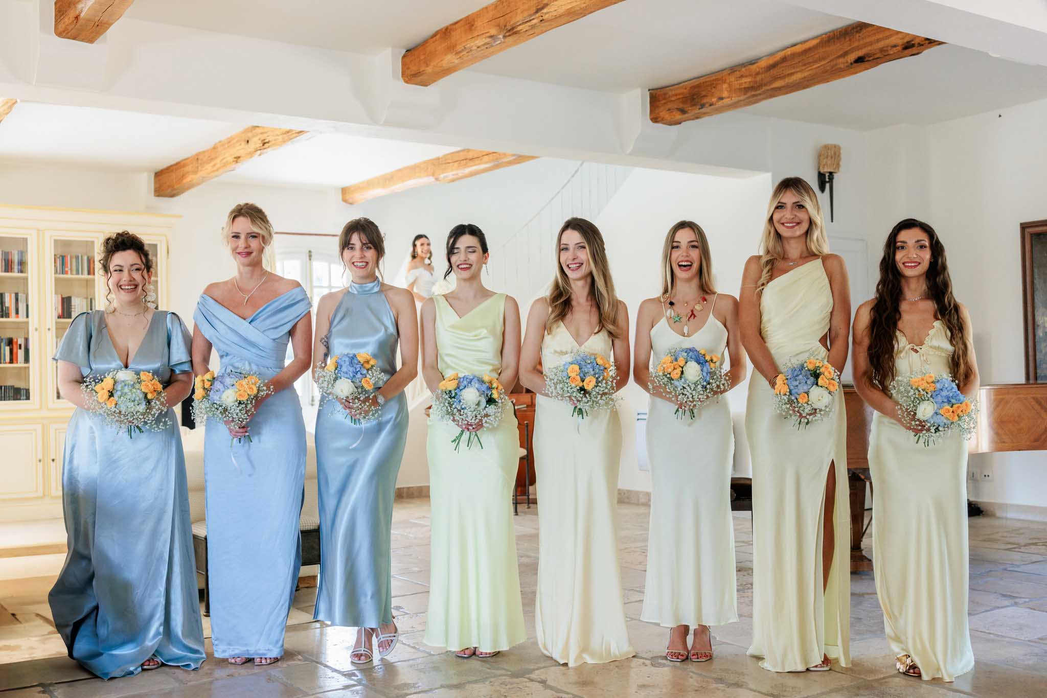Eight bridesmaids in dusty blue and sage gowns holding matching bouquets of blue delphinium and yellow ranunculus