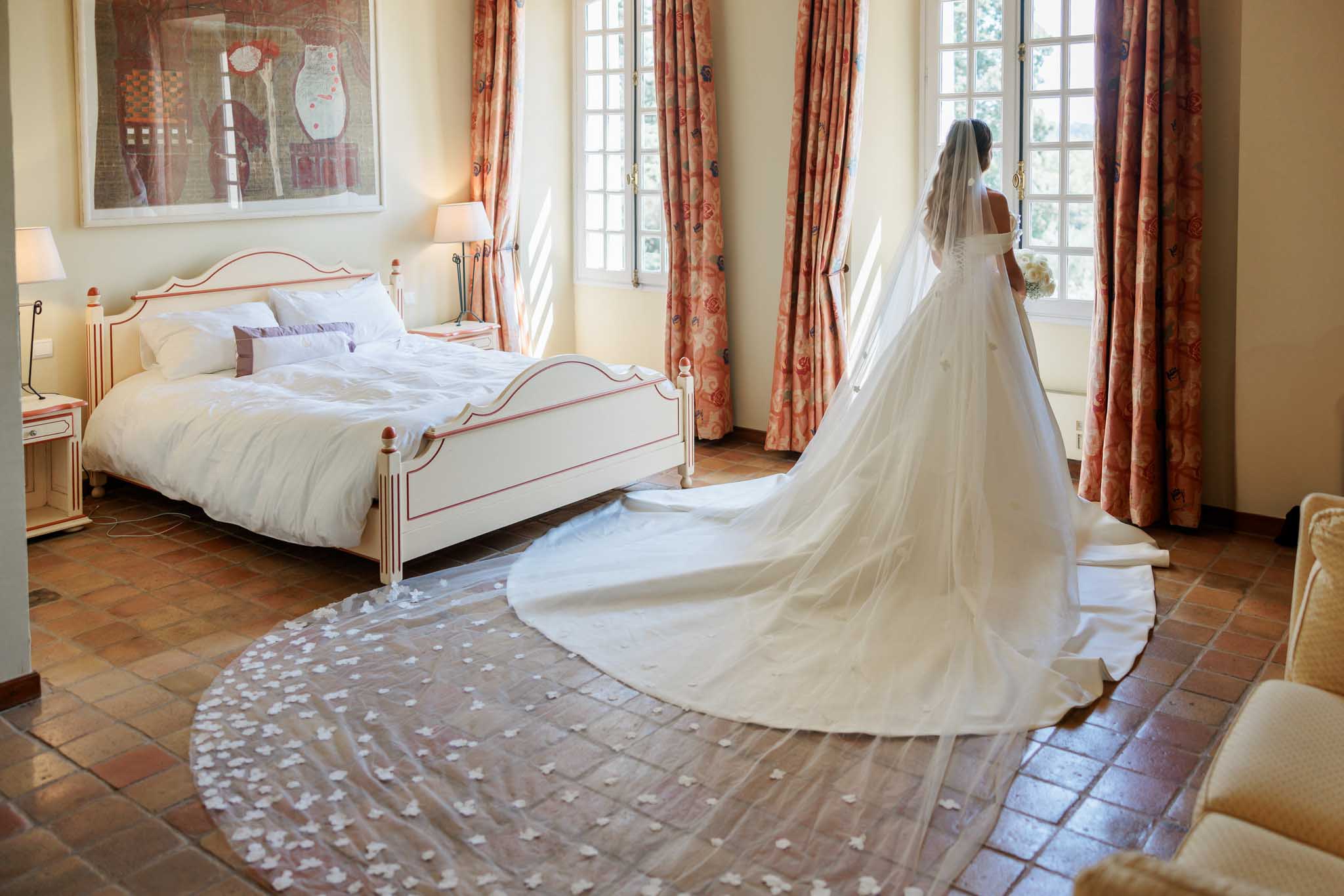 Bride in full-length gown and cathedral train standing in a classical bedroom with petals scattered on floor