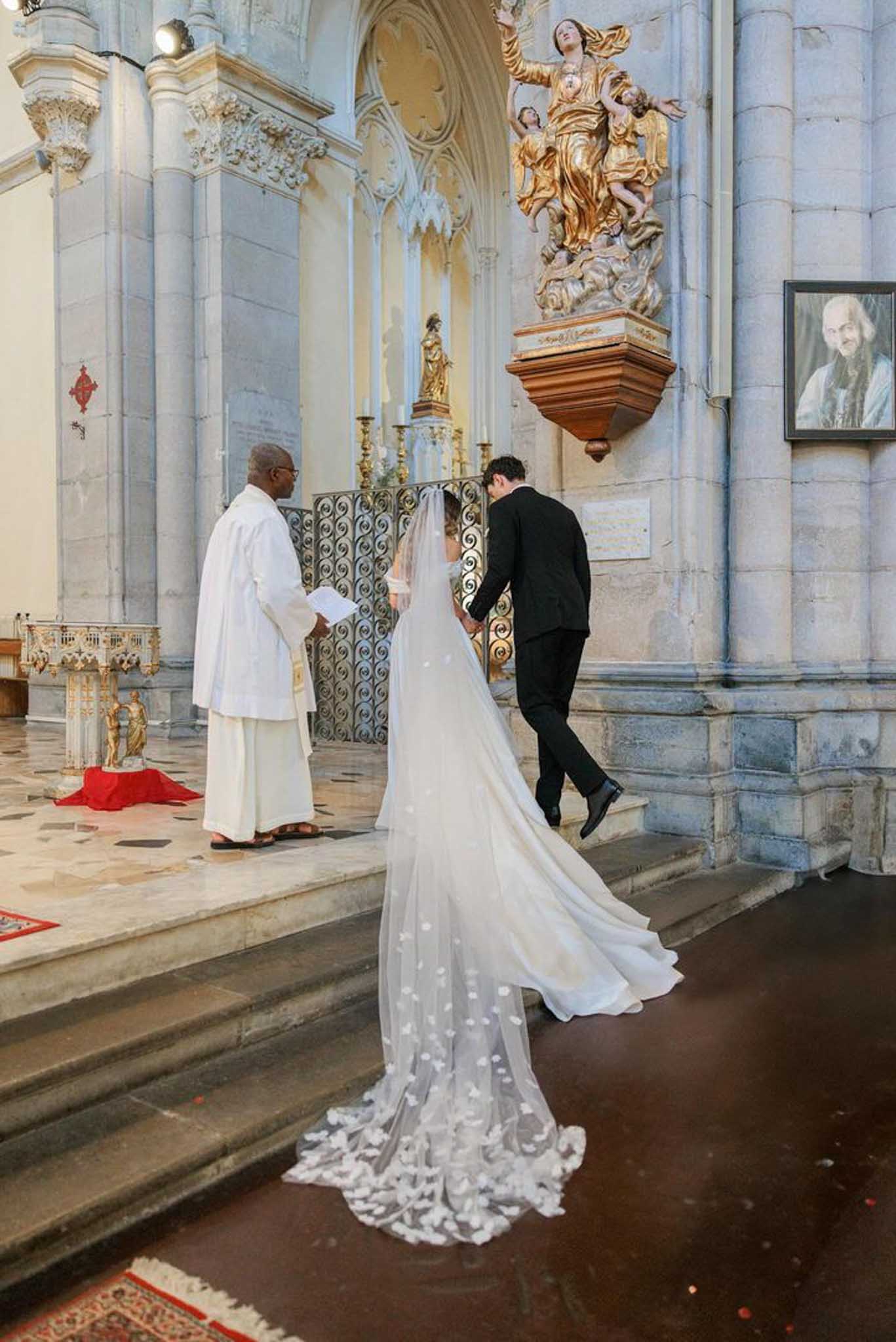 Bride in ivory column gown walks toward groom at ornate gold-leaf chapel altar with stone columns and pulpit