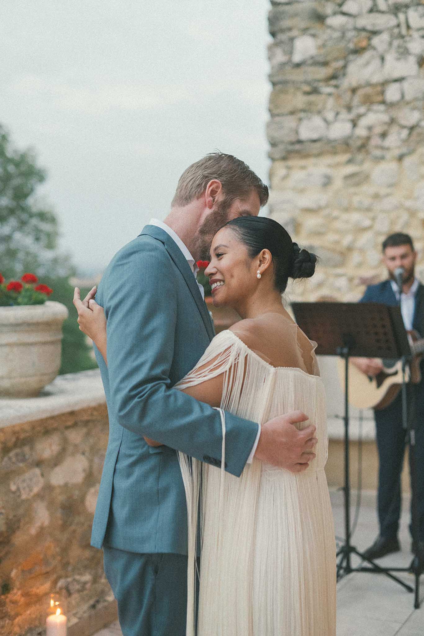 Bride and groom sharing first dance at outdoor reception against historic stone building with red geraniums