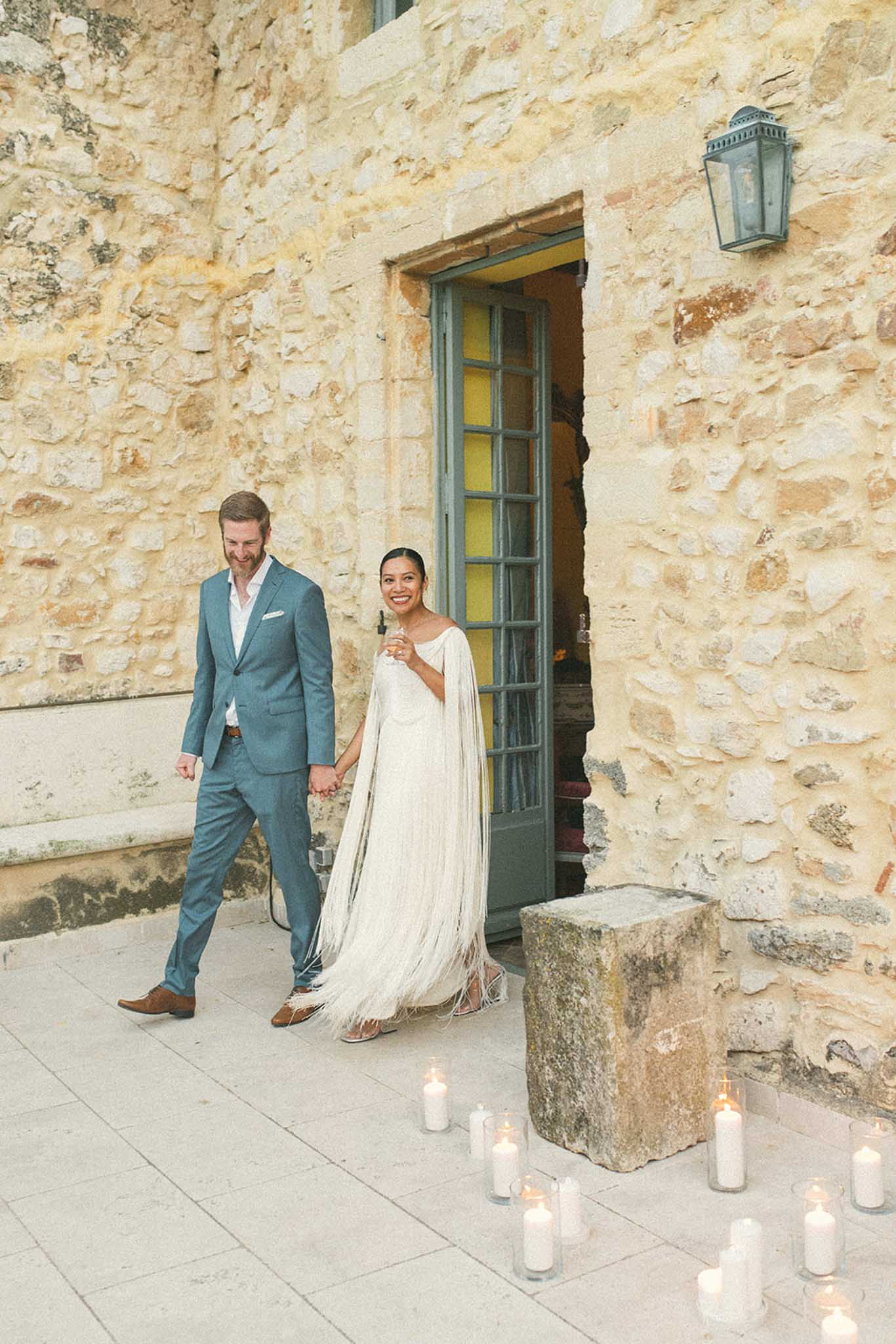 Couple walking hand-in-hand through stone courtyard, bride in ivory fringe cape dress, groom in teal suit, candles on ground