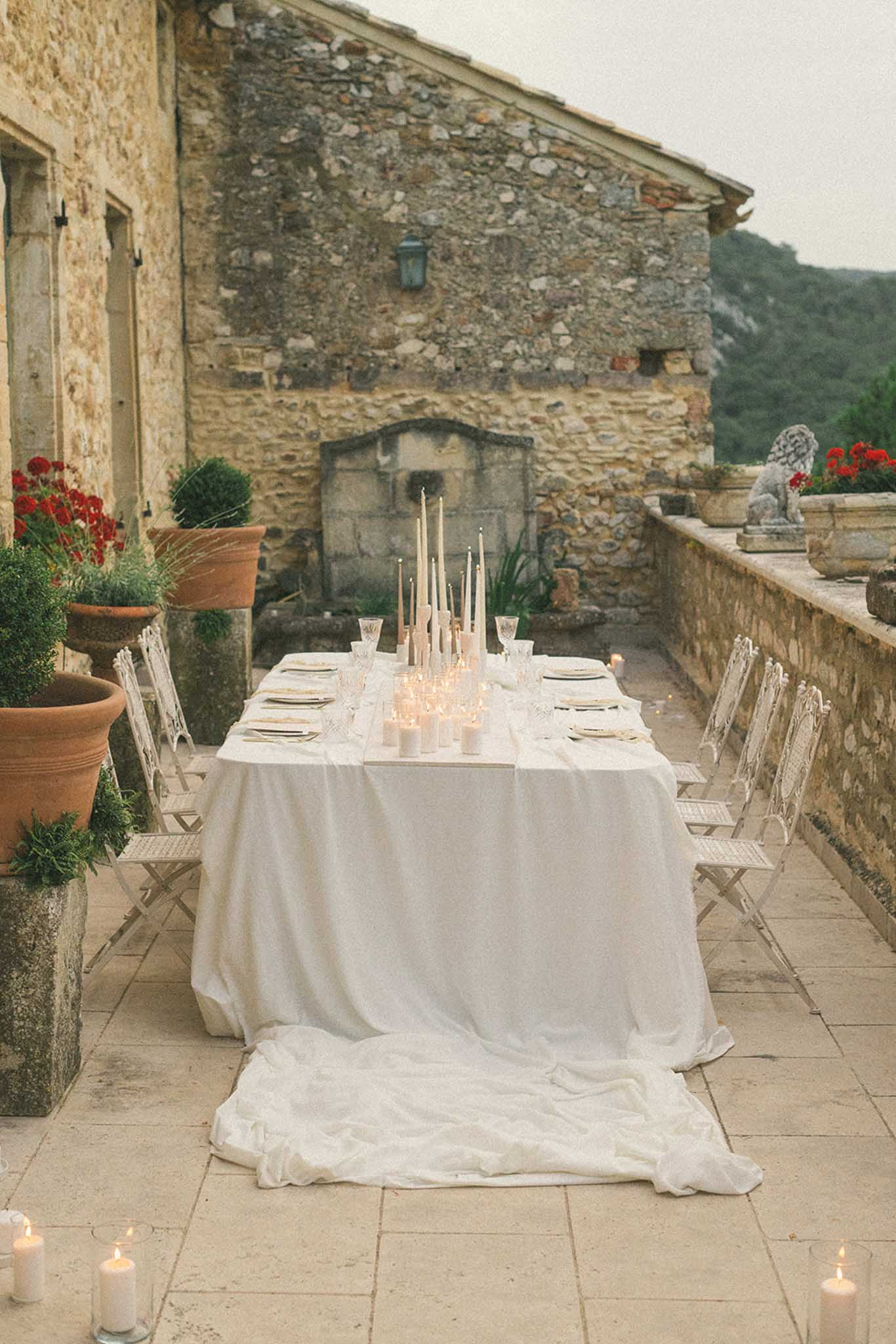 Reception table on stone terrace with ivory linen, cream taper candles in glass holders, and terracotta pots with red flowers