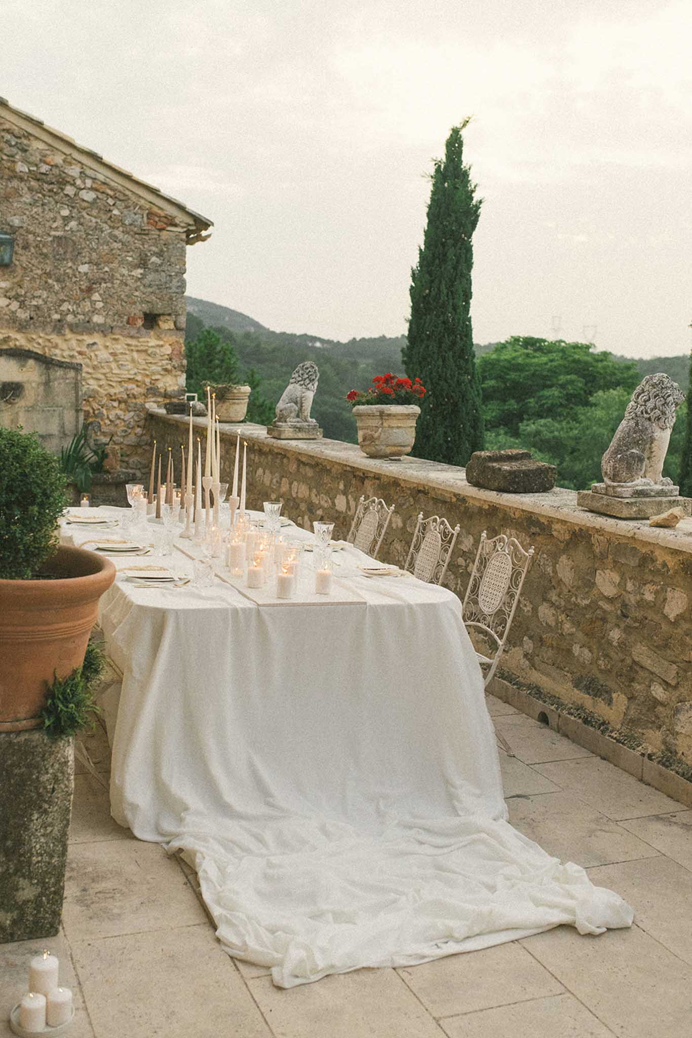 Single round table on stone terrace with white linen, candles, and rattan chairs overlooking forested hills