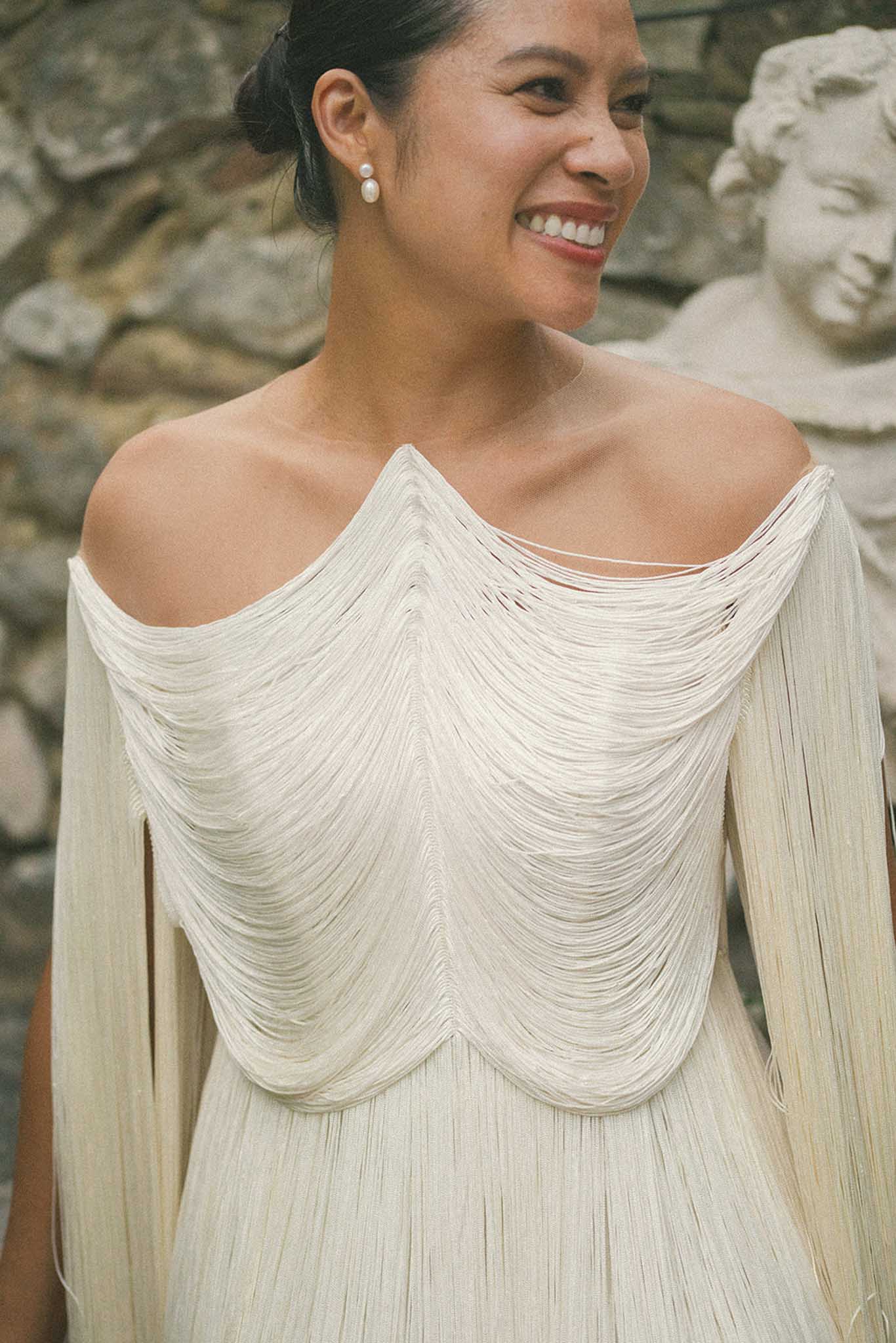 Smiling bride in off-shoulder pleated ivory wedding dress with pearl earrings against stone backdrop