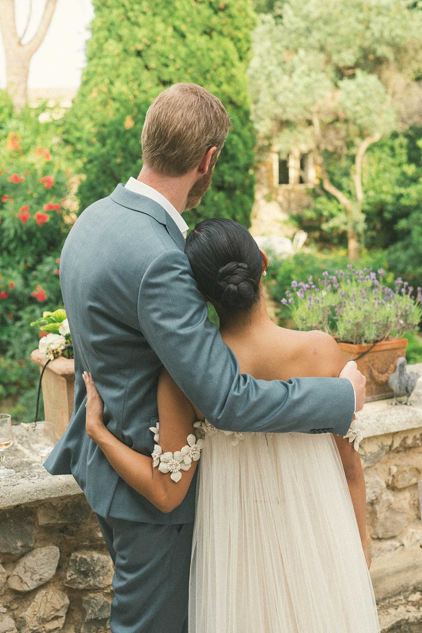 Groom in slate blue suit carrying bride through Mediterranean garden with ivy walls and terracotta planters