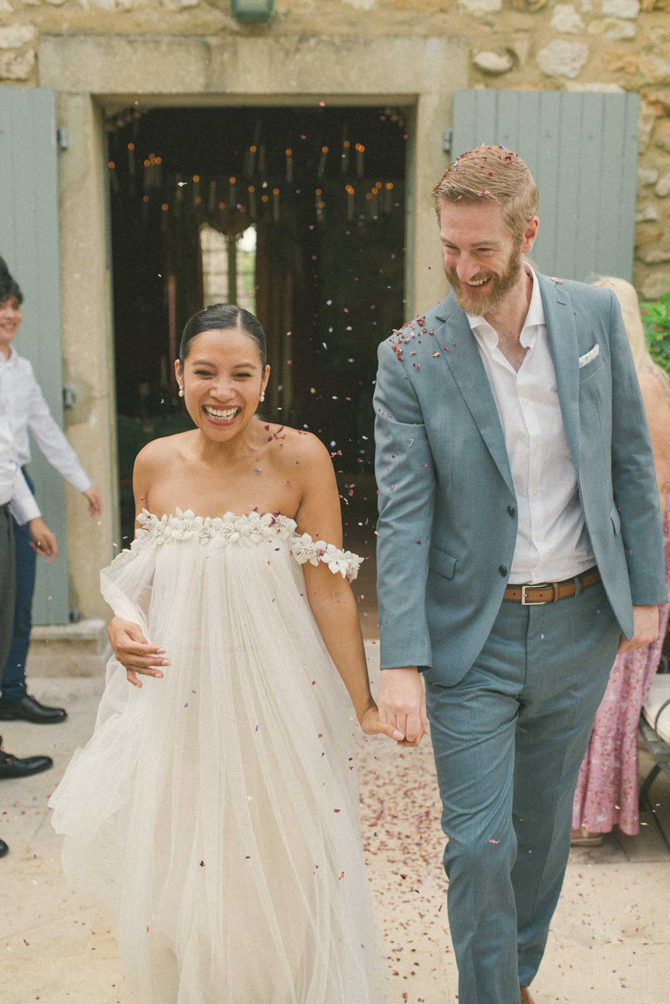 Bride and groom walking through stone courtyard exit as guests throw red and gold confetti, blue shutters in background