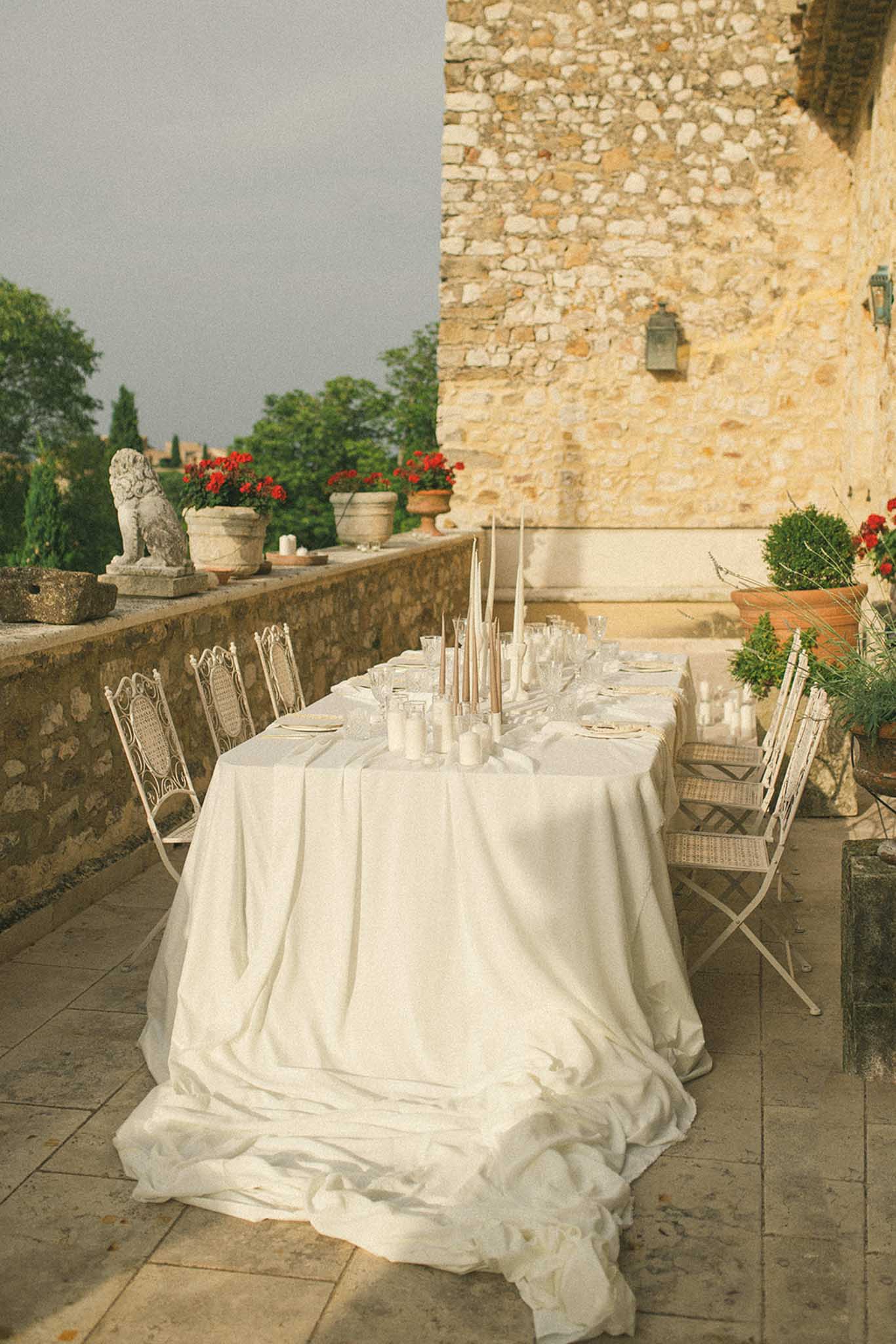 Reception table on stone terrace of historic château with rattan chairs, red geraniums and lion statue in background