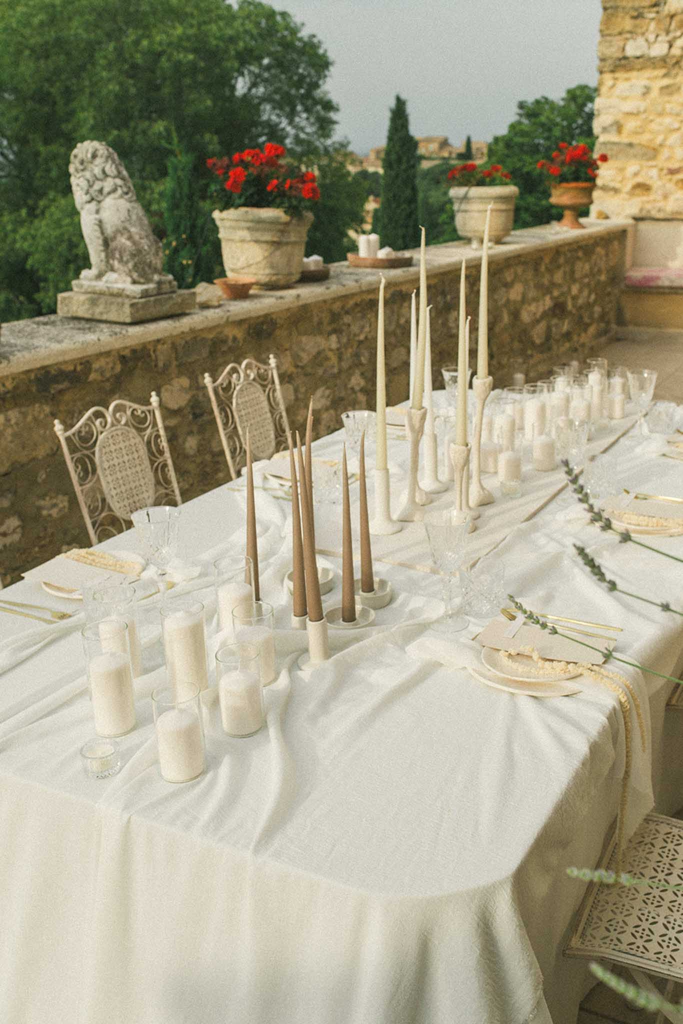 Long dining table on a stone terrace with ivory candles, taper candles and eucalyptus; red geraniums on the balustrade behind.
