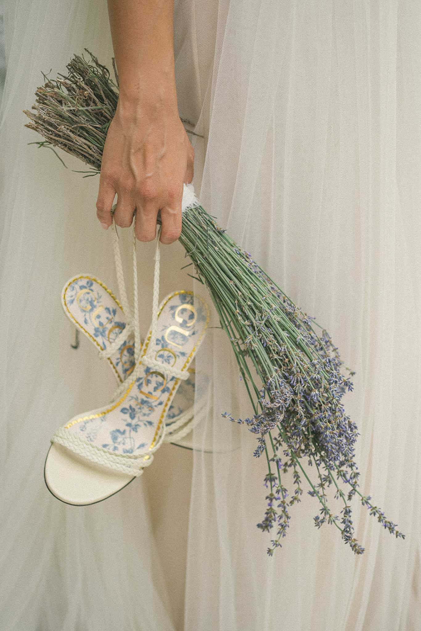 A close-up detail shot of a bride's hand holding two items simultaneously against the backdrop of a sheer ivory tulle veil or skirt. In one hand she carries a pair of Gucci heeled sandals featuring a white and blue floral toile print insole with gold logo lettering and cream rope-style ankle straps, and in the other she holds a loosely bundled bouquet of fresh lavender with long green stems and purple blooms. The pairing of the designer shoes with the unstructured lavender bundle creates a deliberate contrast between polished bridal accessories and a relaxed, Provençal-inspired floral choice.