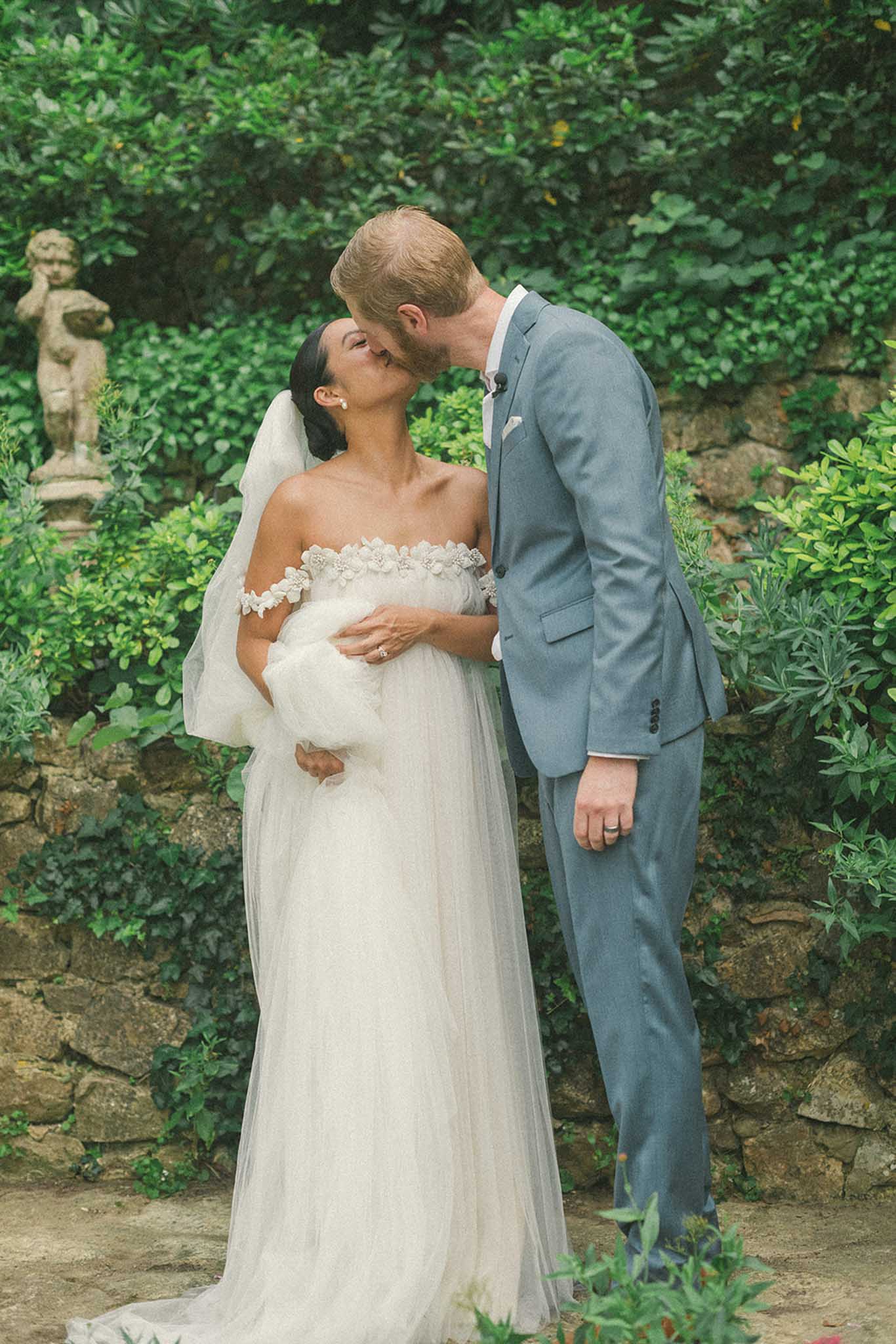 Bride in ivory gown and groom in slate-blue suit kiss before ivy-covered stone walls and classical garden statue