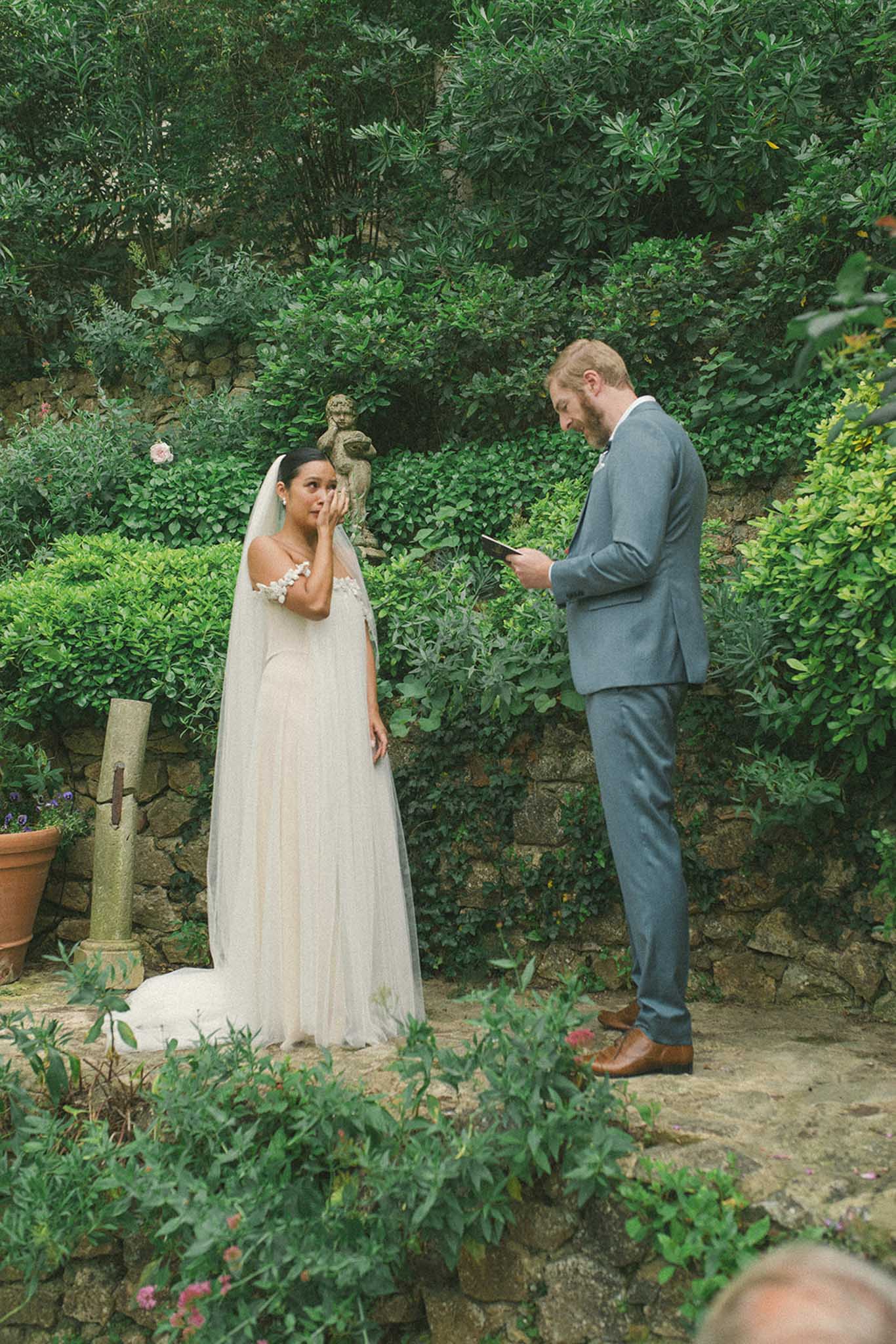 Bride and groom exchanging vows in an ivy-covered stone garden, groom in slate-blue suit reading from a card