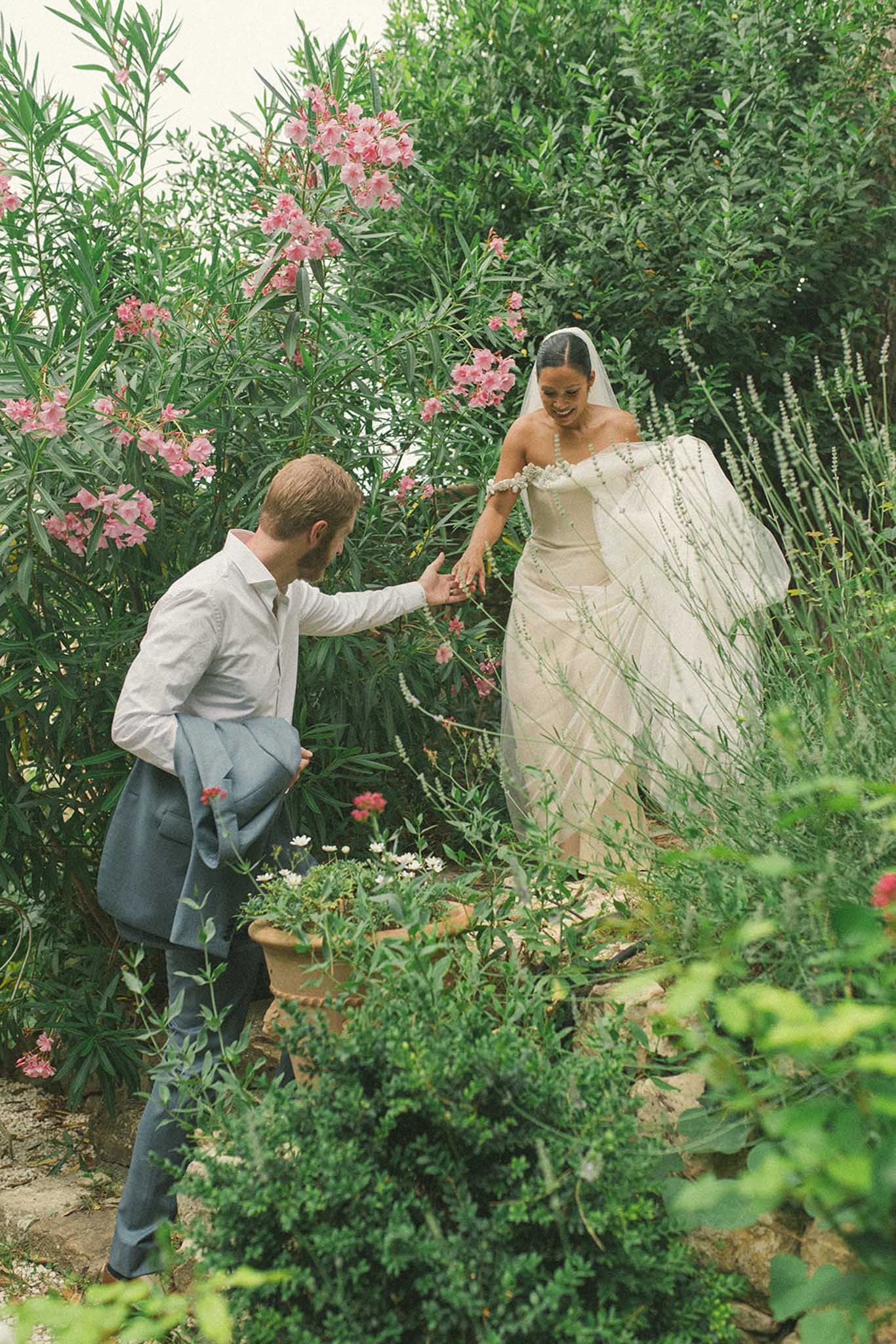 Bride and groom walking together through Mediterranean herb garden, groom in pale blue suit, bride in ivory off-shoulder gown