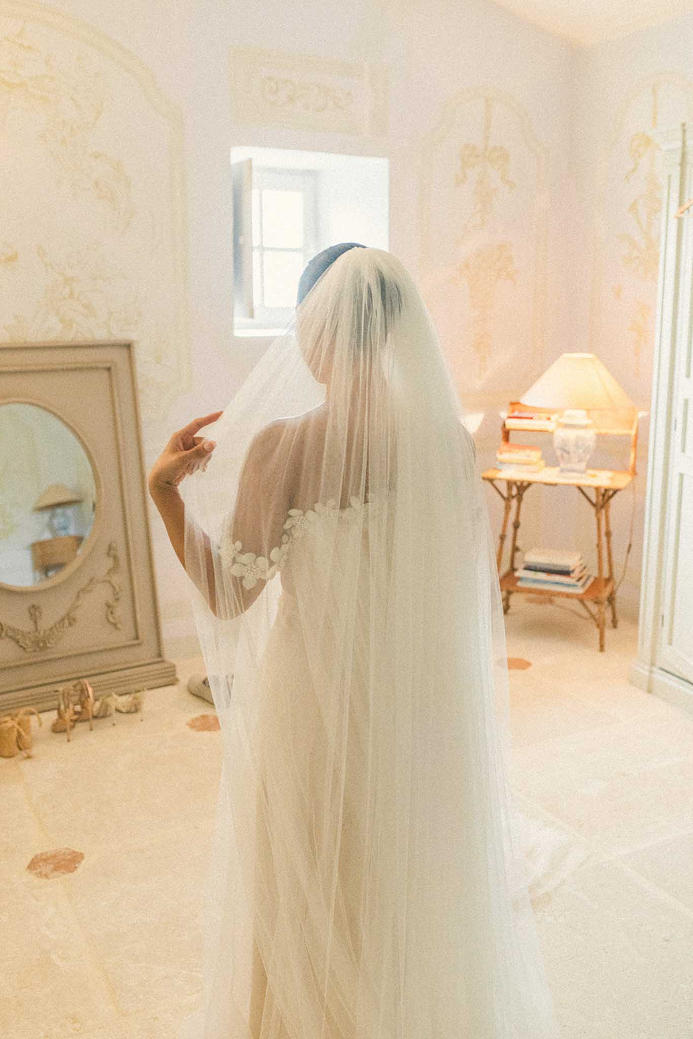 Bride standing with flowing veil and blue headpiece in elegant bedroom with damask wallpaper