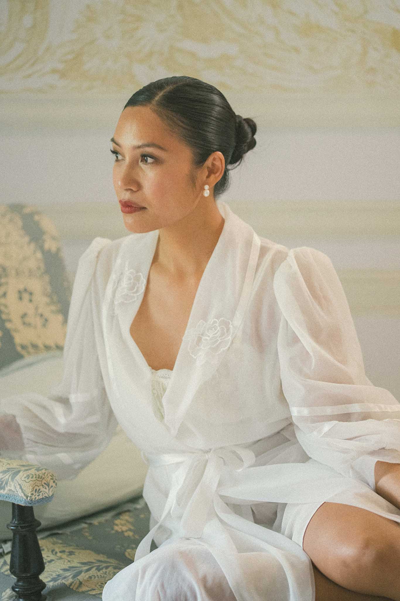 Bride seated in white silk robe with embroidered roses gazing to the side in a classical interior