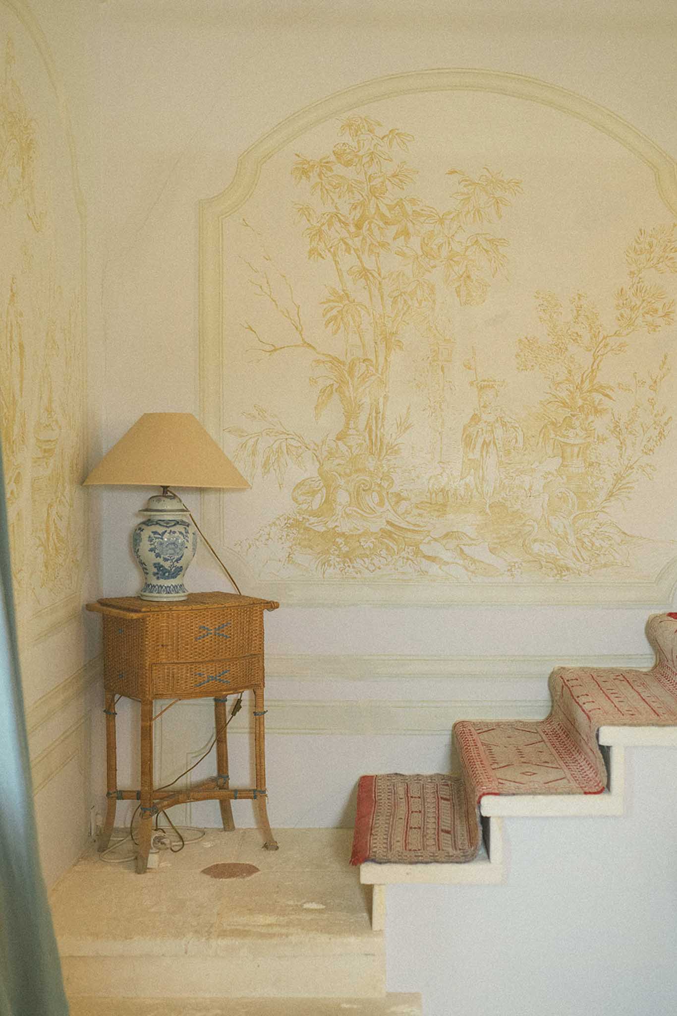 Bridal suite corner with Chinoiserie botanical mural, rattan side table, blue porcelain lamp, and patterned textiles